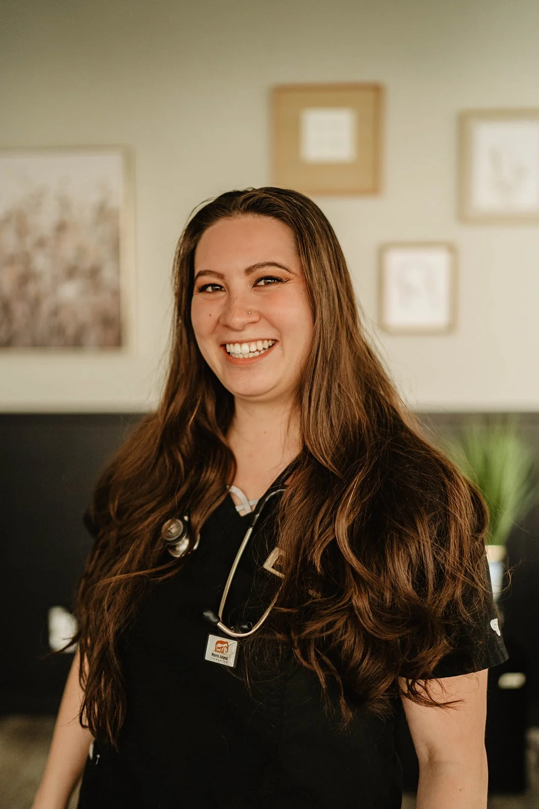 Smiling person with long hair wearing a stethoscope and black scrubs, standing indoors with framed artwork on the wall behind.