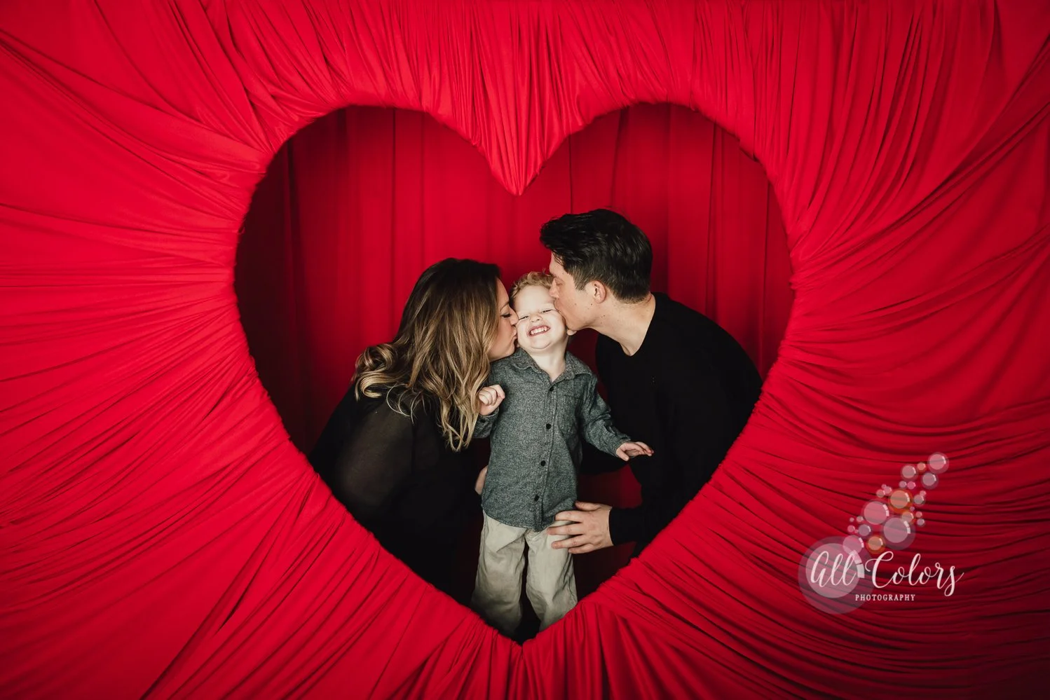 family of three behind a red fabric draped heart backdrop