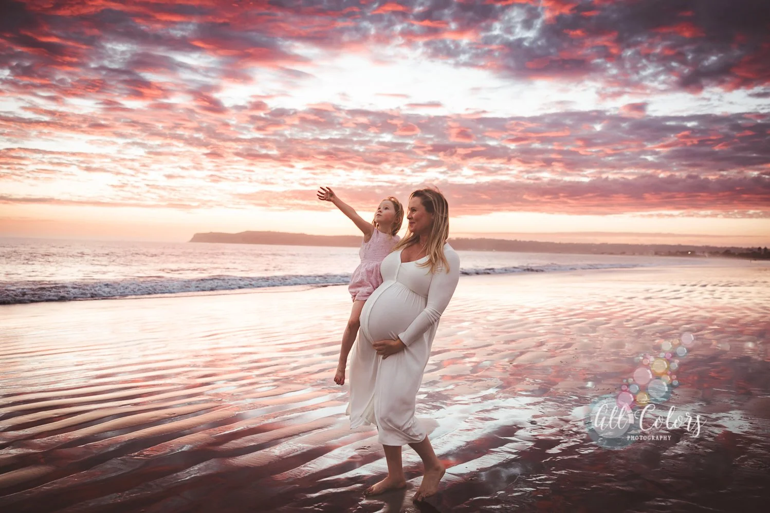 Expecting woman wearing a white dress plays with young daughter at the beach at sunset