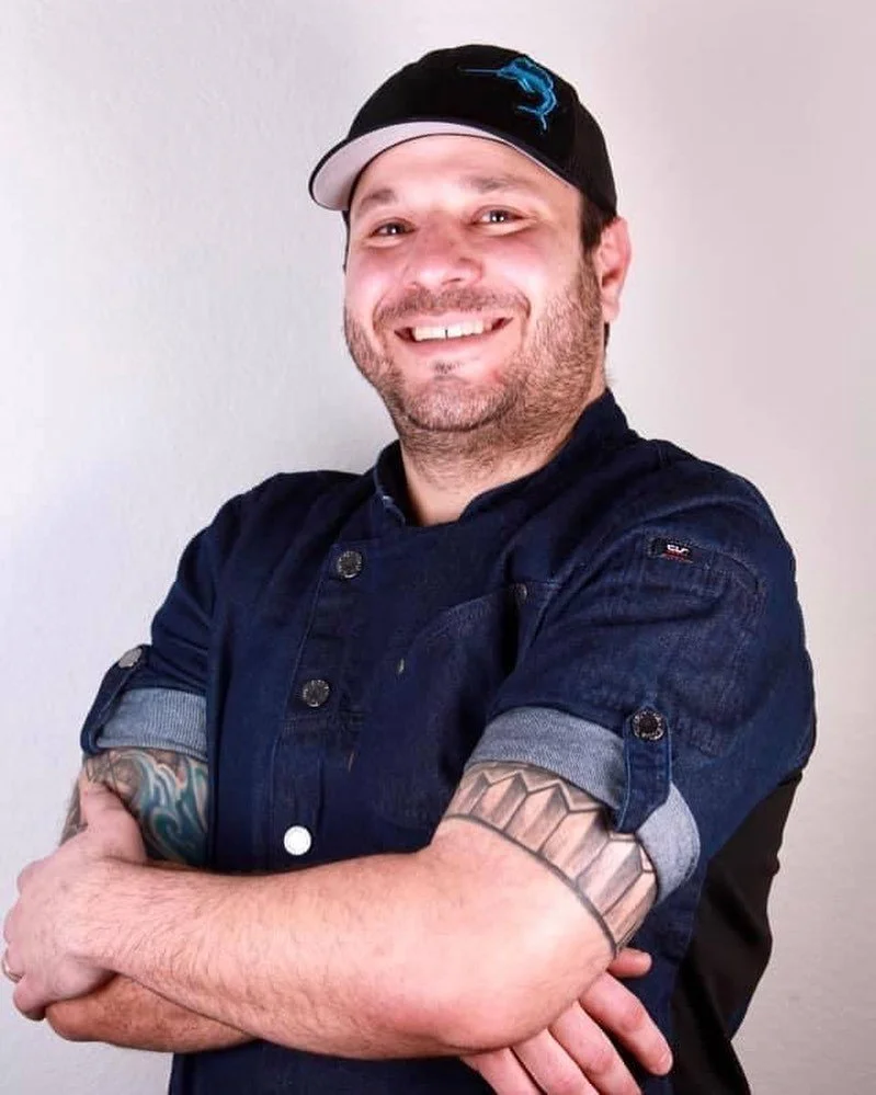 Man wearing a black baseball cap with a blue logo, a dark denim chef coat with rolled-up sleeves, smiling, with tattoos on his arms, standing against a plain light gray background.