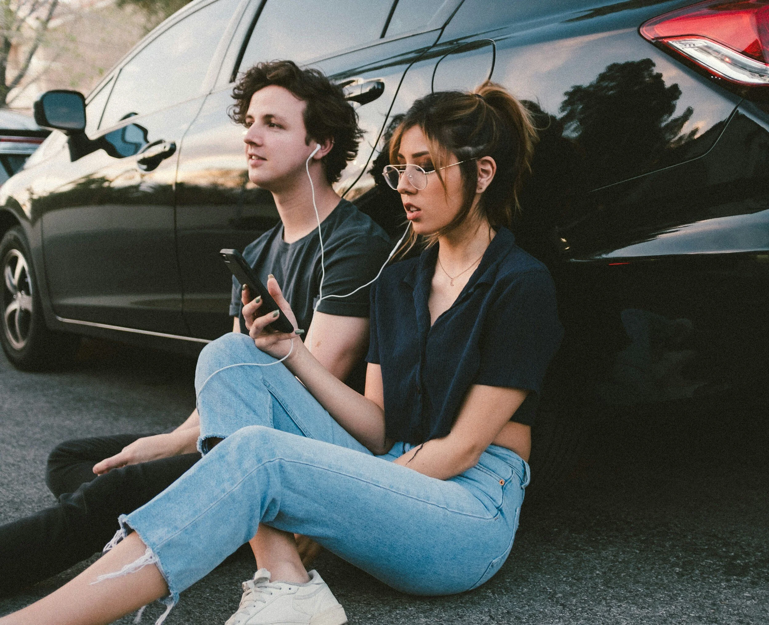 Cool couple sat on the floor leaning against a car sharing headphones and listening to a podcast.