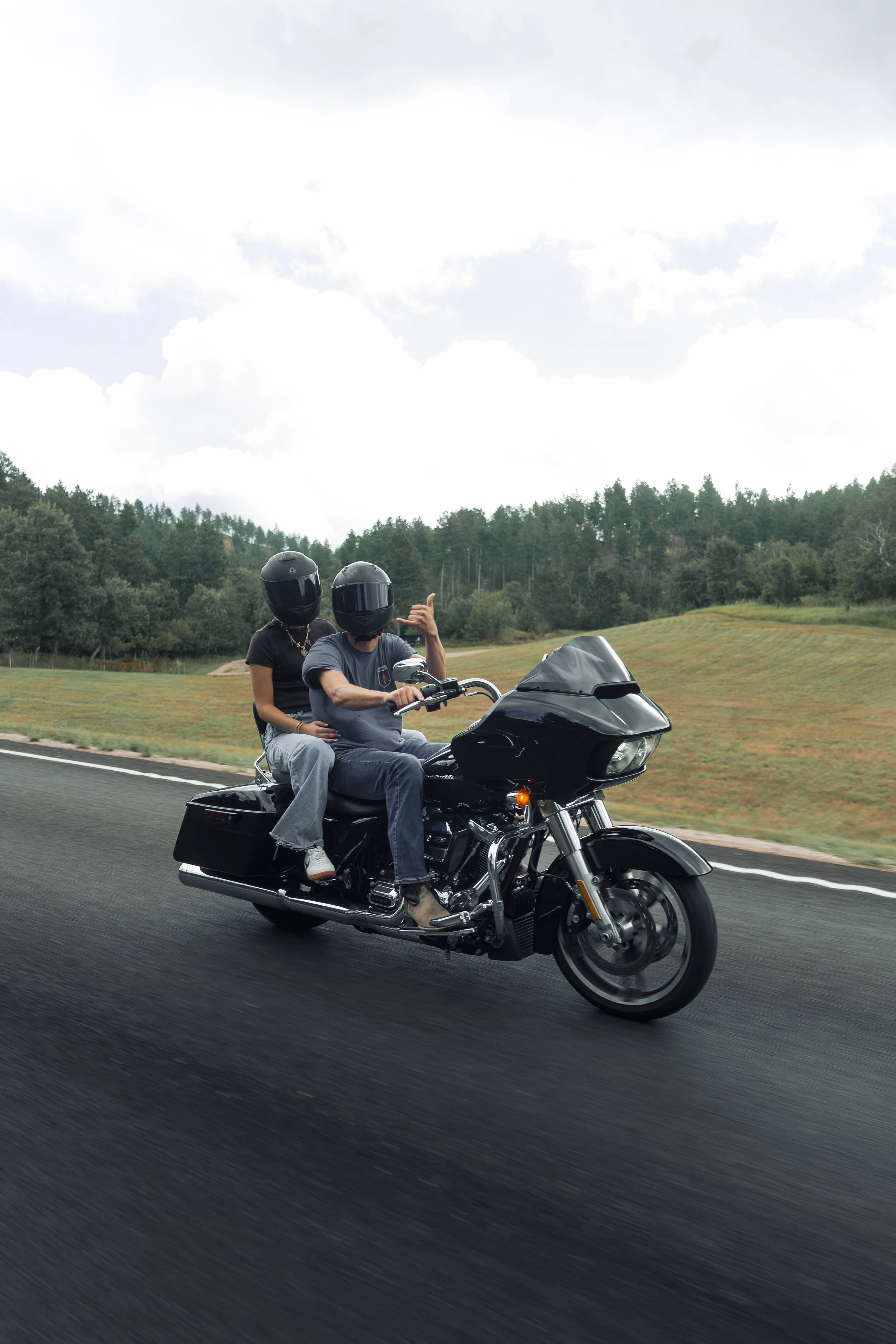 Two people wearing black helmets riding a black motorcycle on a highway, with trees and cloudy sky in the background.