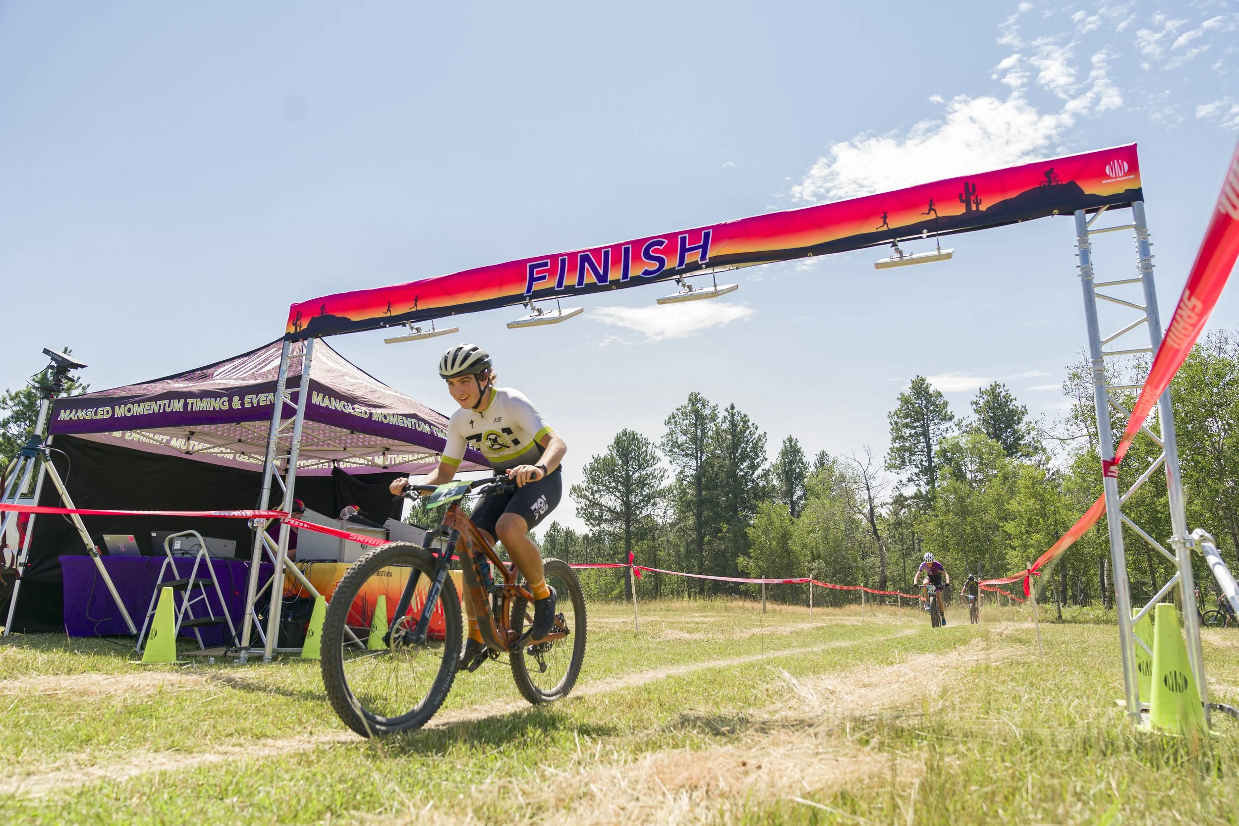 A person riding a mountain bike crosses a finish line at an outdoor race. The scene is set on a grassy trail with trees in the background and a bright, sunny sky.
