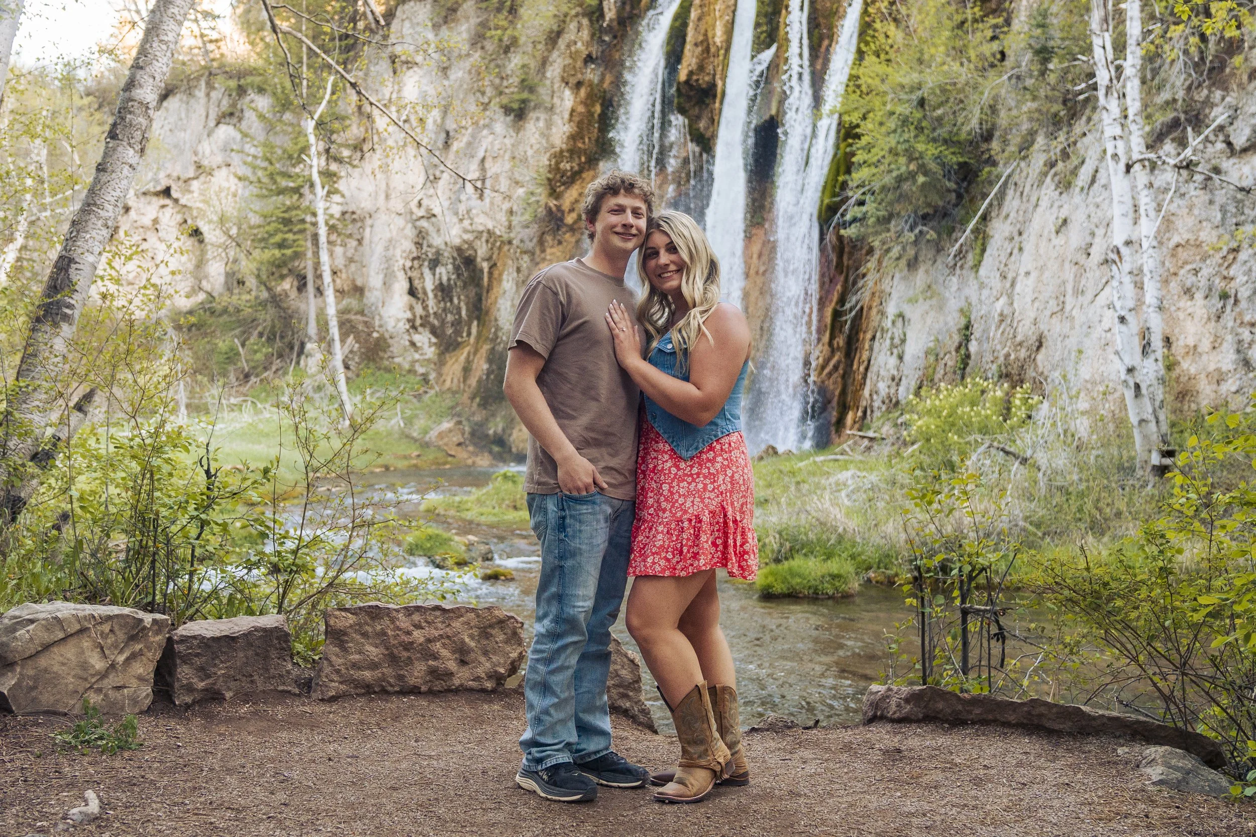 A happy couple standing arm in arm near a creek with a waterfall and trees in the background.