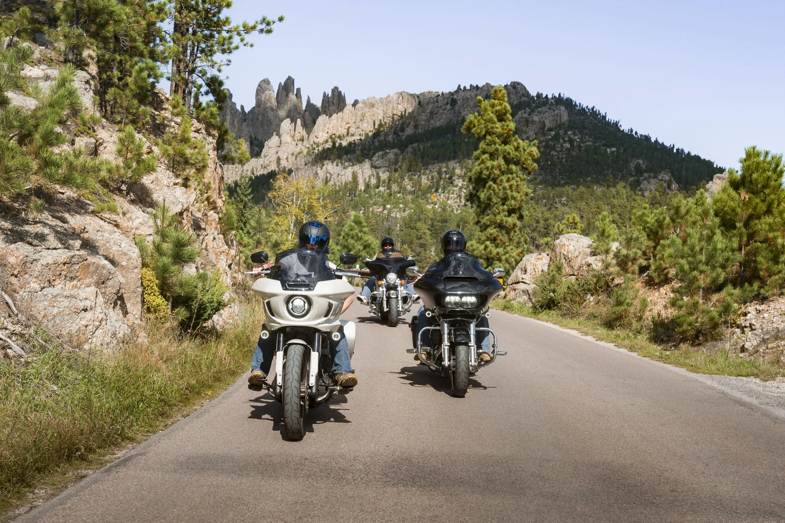 Three motorcyclists riding on a mountain road surrounded by trees and rocky peaks in the background.
