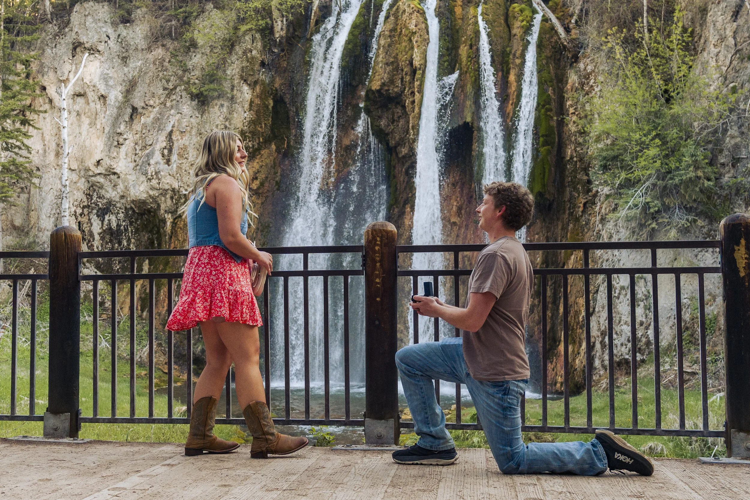 A man on one knee proposes to a woman by a waterfall at a scenic outdoor location, with a wooden fence and green trees around them.