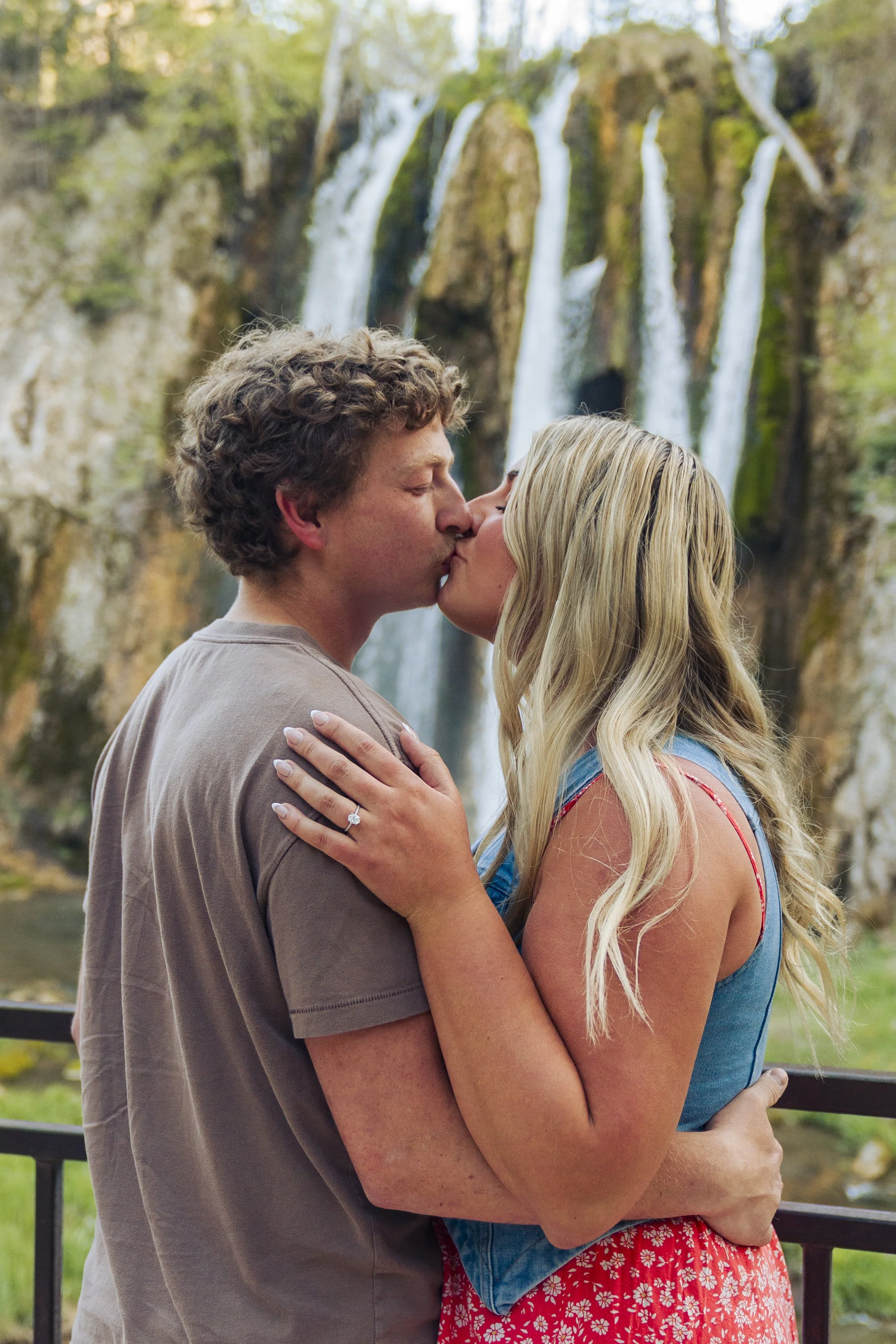 A couple sharing a kiss outdoors with a waterfall in the background.