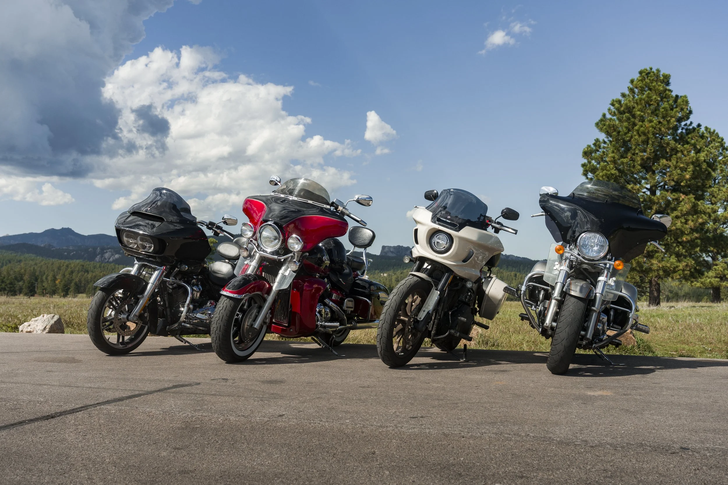 Four motorcycles parked on a paved area with a scenic background of green trees, mountains, and a bright blue sky with a few clouds.