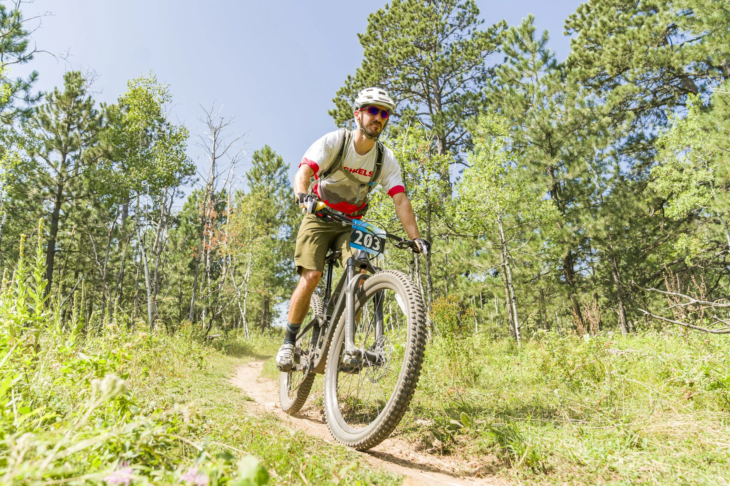 A man mountain biking on a trail through a green forest during the daytime. He is wearing a helmet, sunglasses, and casual biking attire, and riding a black mountain bike with a racing number 203.