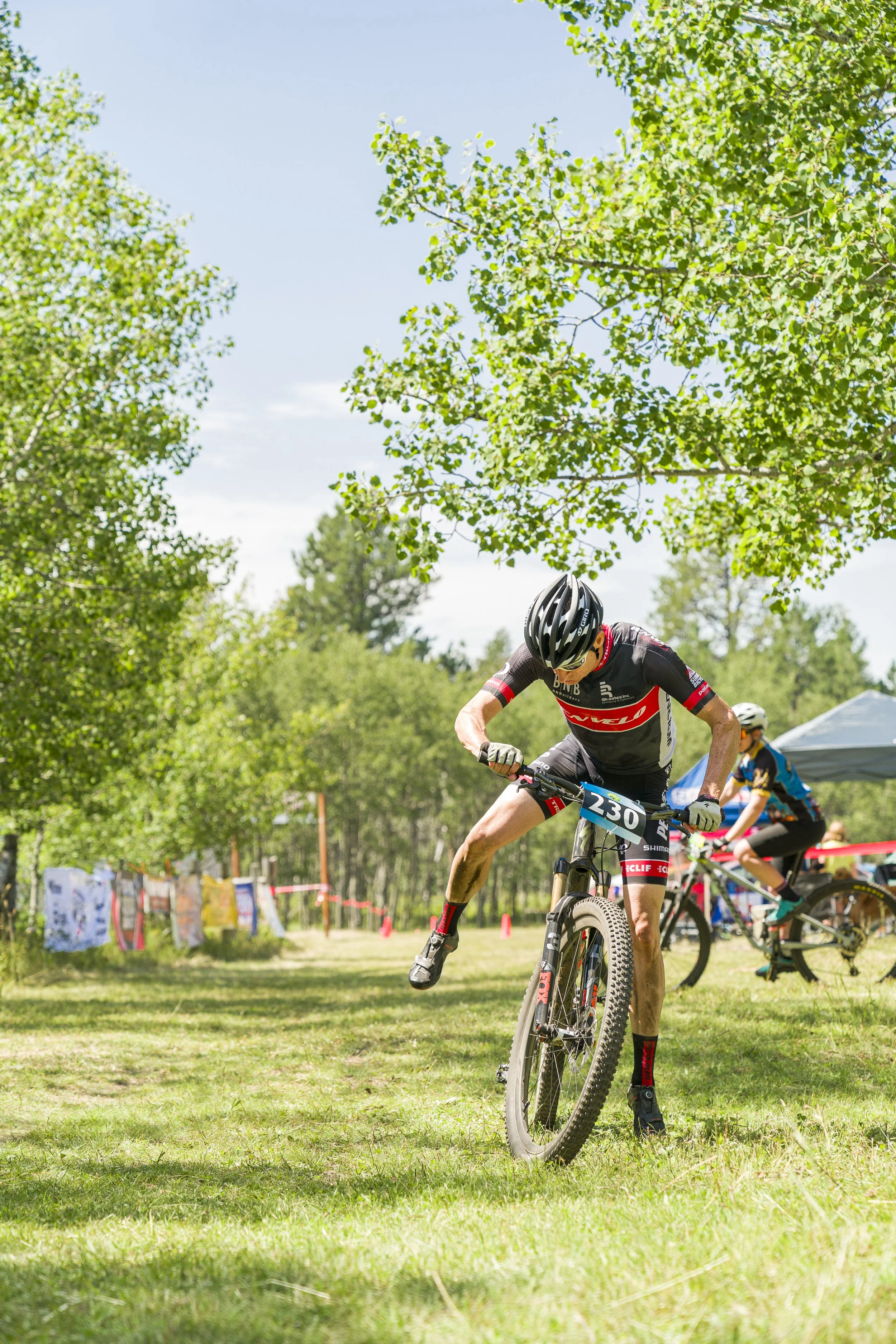 A mountain biker in a black and red jersey and helmet riding on a grassy trail, with trees and other bikers in the background during a race.
