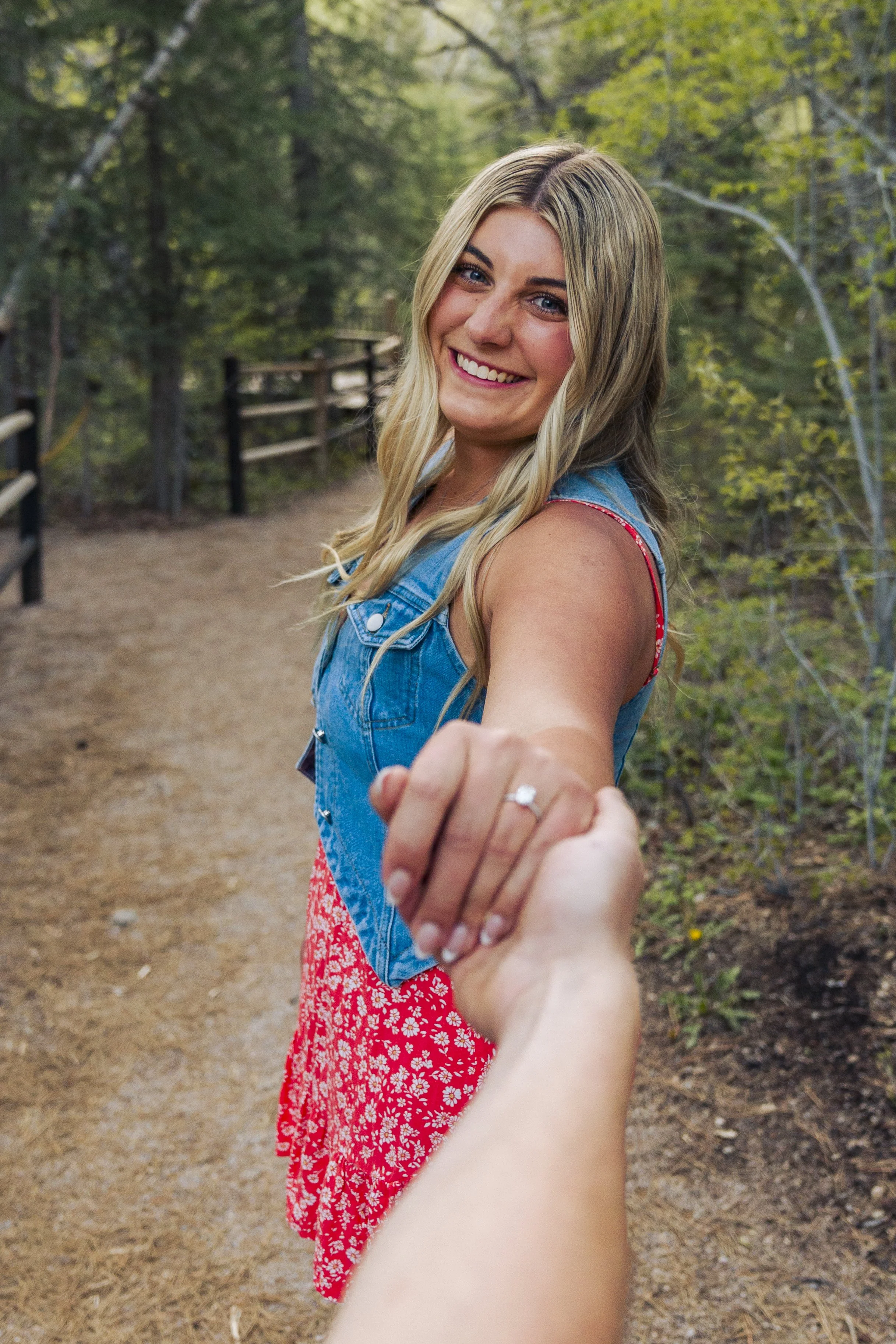A young woman with long blonde hair smiling and holding hands with the person taking the photo. She is outdoors on a forest trail, wearing a red floral dress and a denim vest.