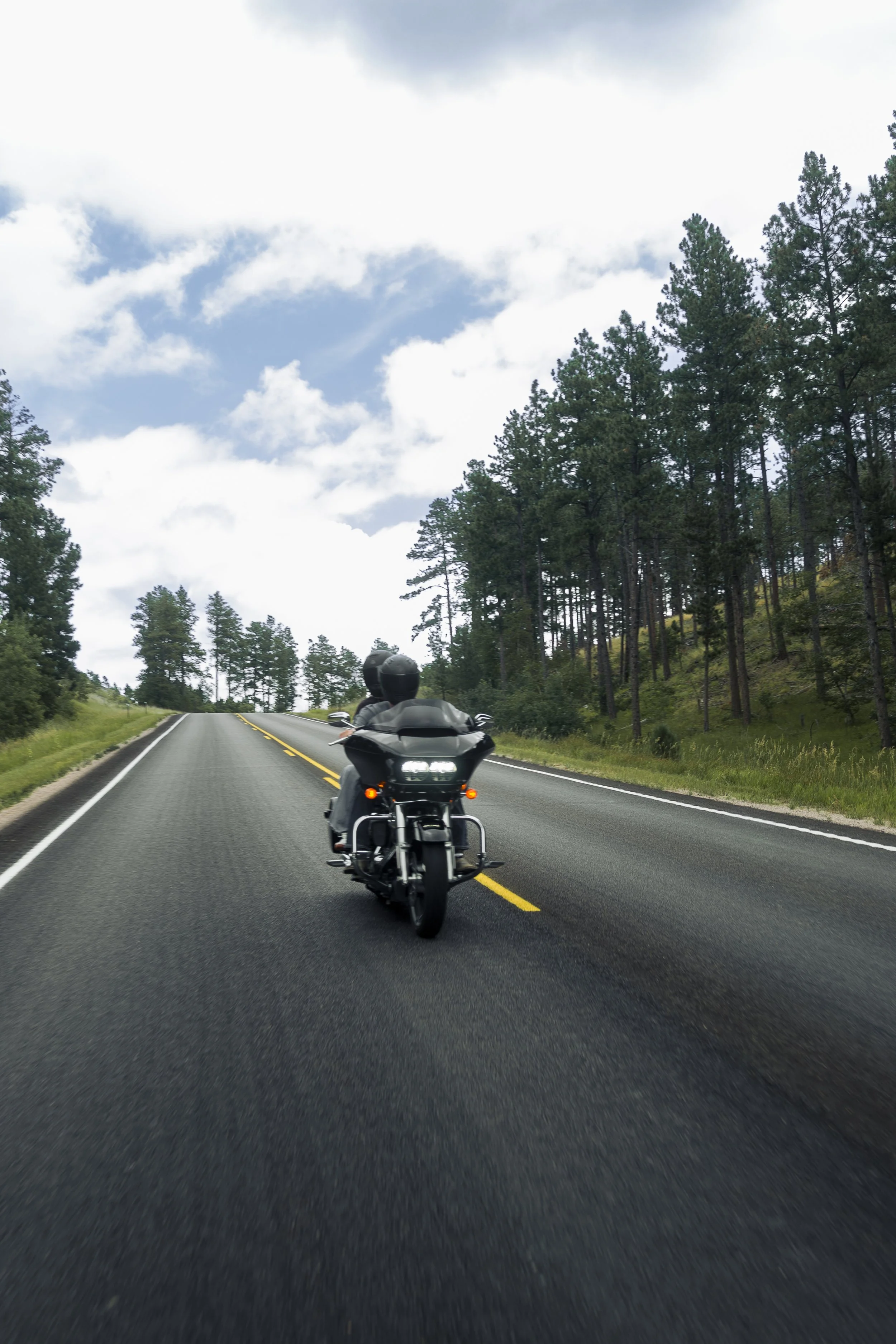 Motorcycle with two riders riding on a two-lane road through a forested area under a partly cloudy sky.