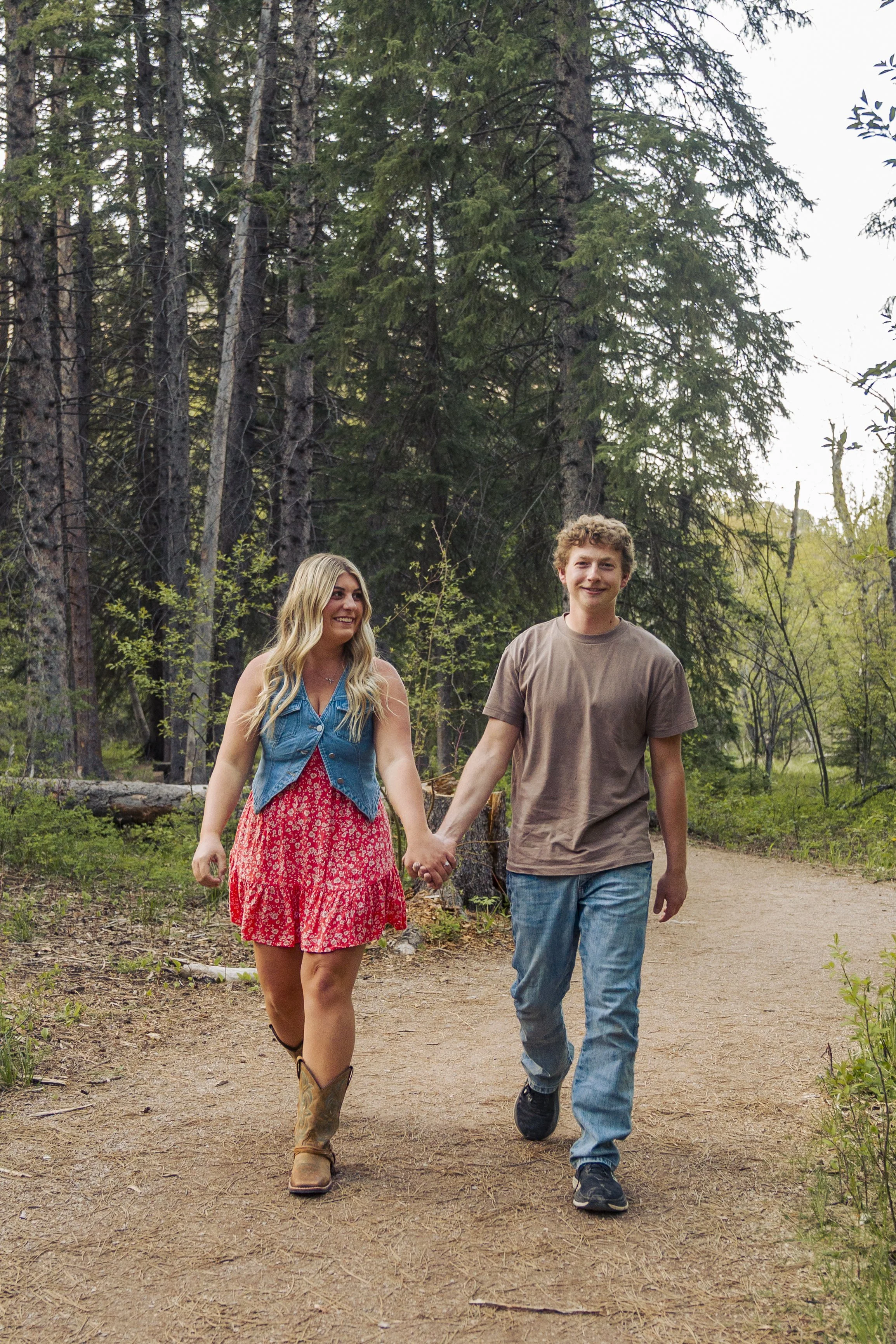 A young couple walking hand in hand along a dirt path in a forest with tall pine trees, the woman wearing a red dress, denim vest, and cowboy boots, and the man wearing a brown t-shirt, jeans, and sneakers, both smiling.