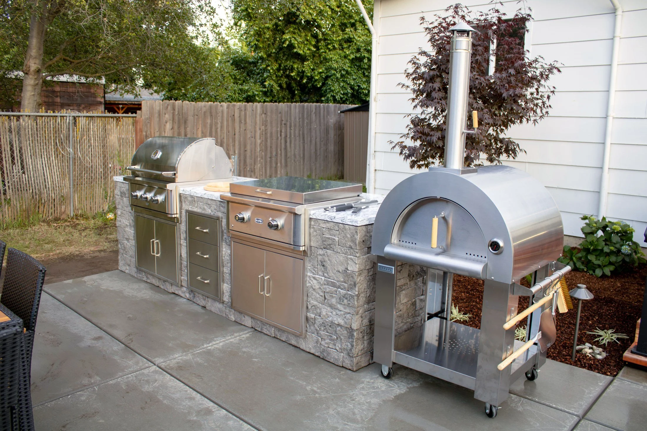 A full outdoor kitchen with a grill, griddle, and pizza oven on a stacked stone base.