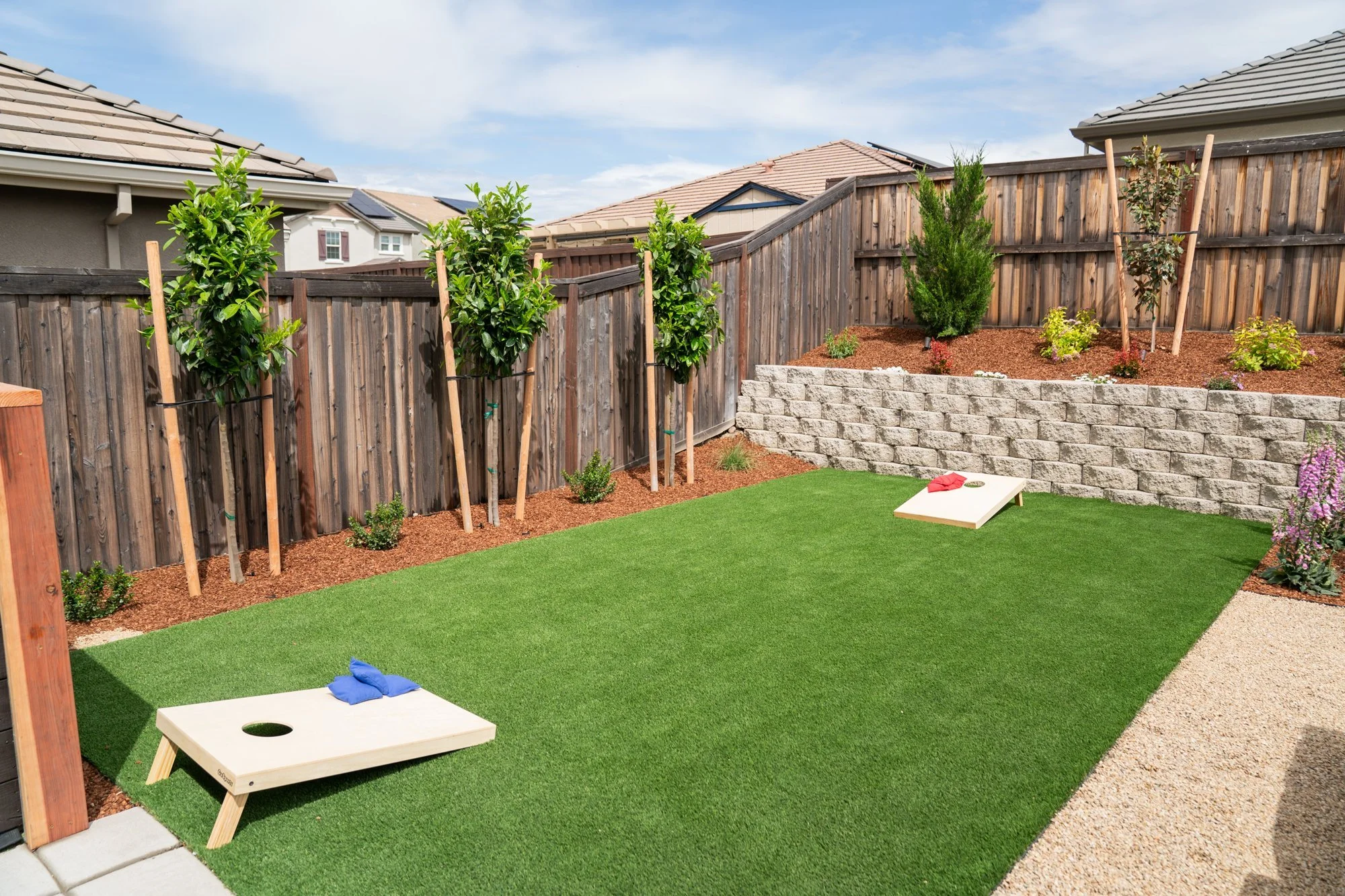 After: synthetic lawn game zone with cornhole boards, retaining wall planting, and young privacy trees - designed for adults, kids, and dogs