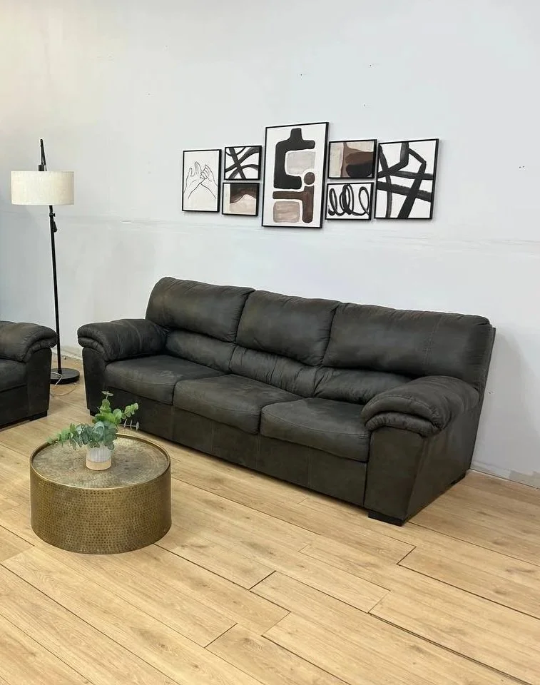 A black leather couch with three seats in a living room, a round metallic coffee table with a small potted plant on it, a white floor lamp, and artwork consisting of six black and white abstract pictures hanging on the wall.