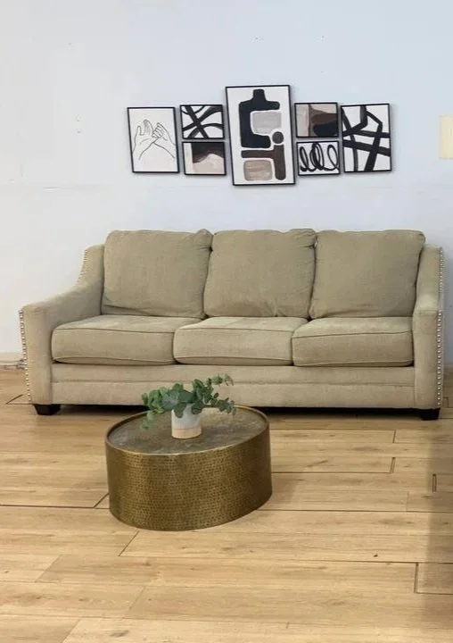 A beige sofa with three cushions, a round textured coffee table with a small potted plant, and abstract black-and-white artwork on the wall behind.