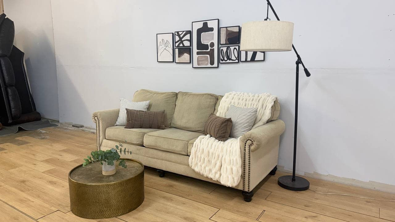A beige sofa with decorative pillows and a white blanket in a living room, with a round gold coffee table holding a small plant and a wall art arrangement above the sofa, and a floor lamp to the right.