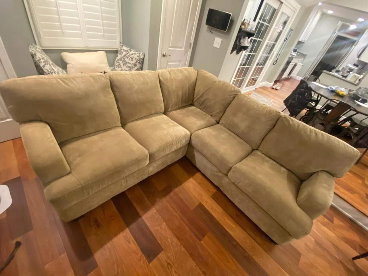 A beige sectional sofa in a living room with wooden floors, kitchen in the background, and a chandelier hanging from the ceiling.