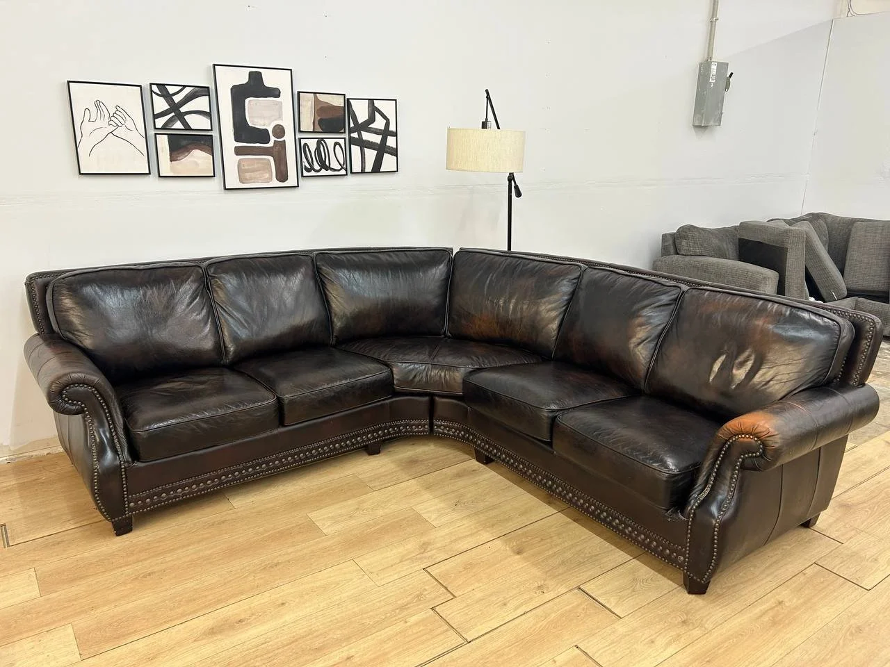 A black leather sectional sofa with nailhead trim on a hardwood floor, with abstract art pieces on the white wall and a standing lamp behind it.