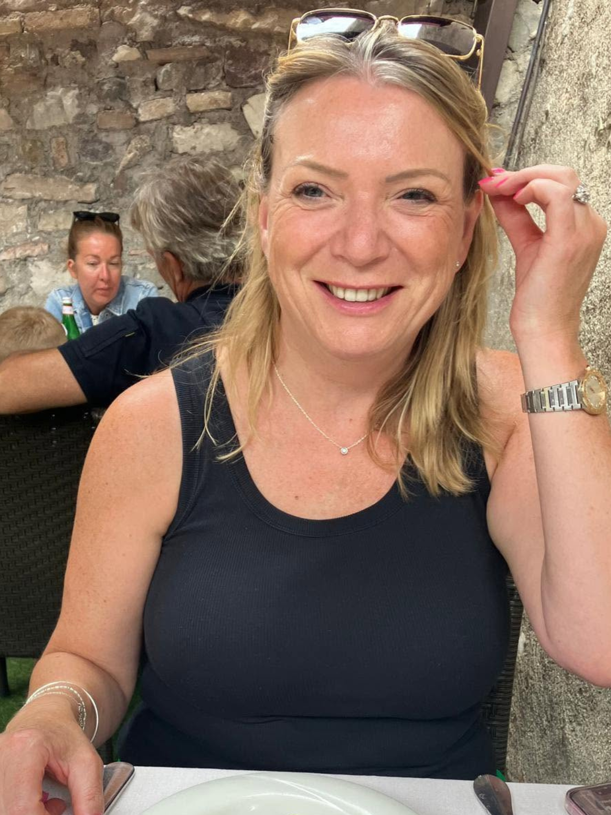 Smiling woman with blond hair, wearing a black sleeveless top, sitting at a table in a restaurant with a stone wall background. She has a pair of sunglasses on her head, a necklace, a watch, and a ring on her finger.