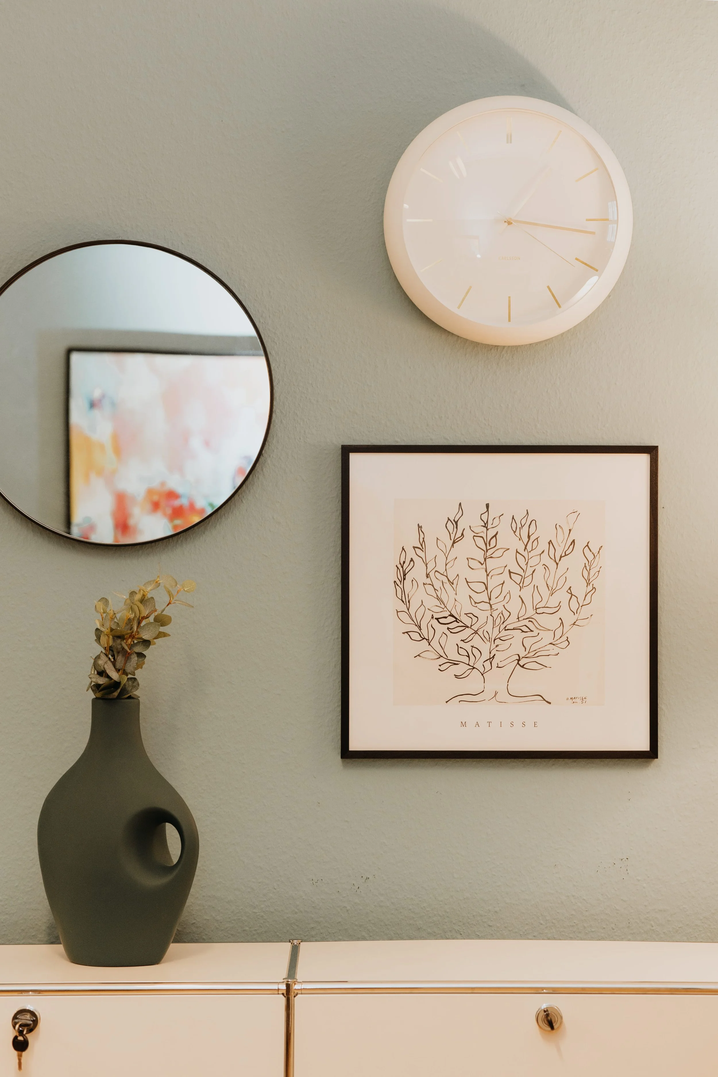 Wand mit drei Dekorationsgegenständen: Ein runder Spiegel, eine weiße Uhr und ein Matisse-Poster mit einer schwarzen Zeichnung eines Baumes. Darunter steht das Wort 'MATISSE'. In der Nähe befindet sich eine graue Vase mit getrockneten Pflanzen auf einem weißen Möbelstück.