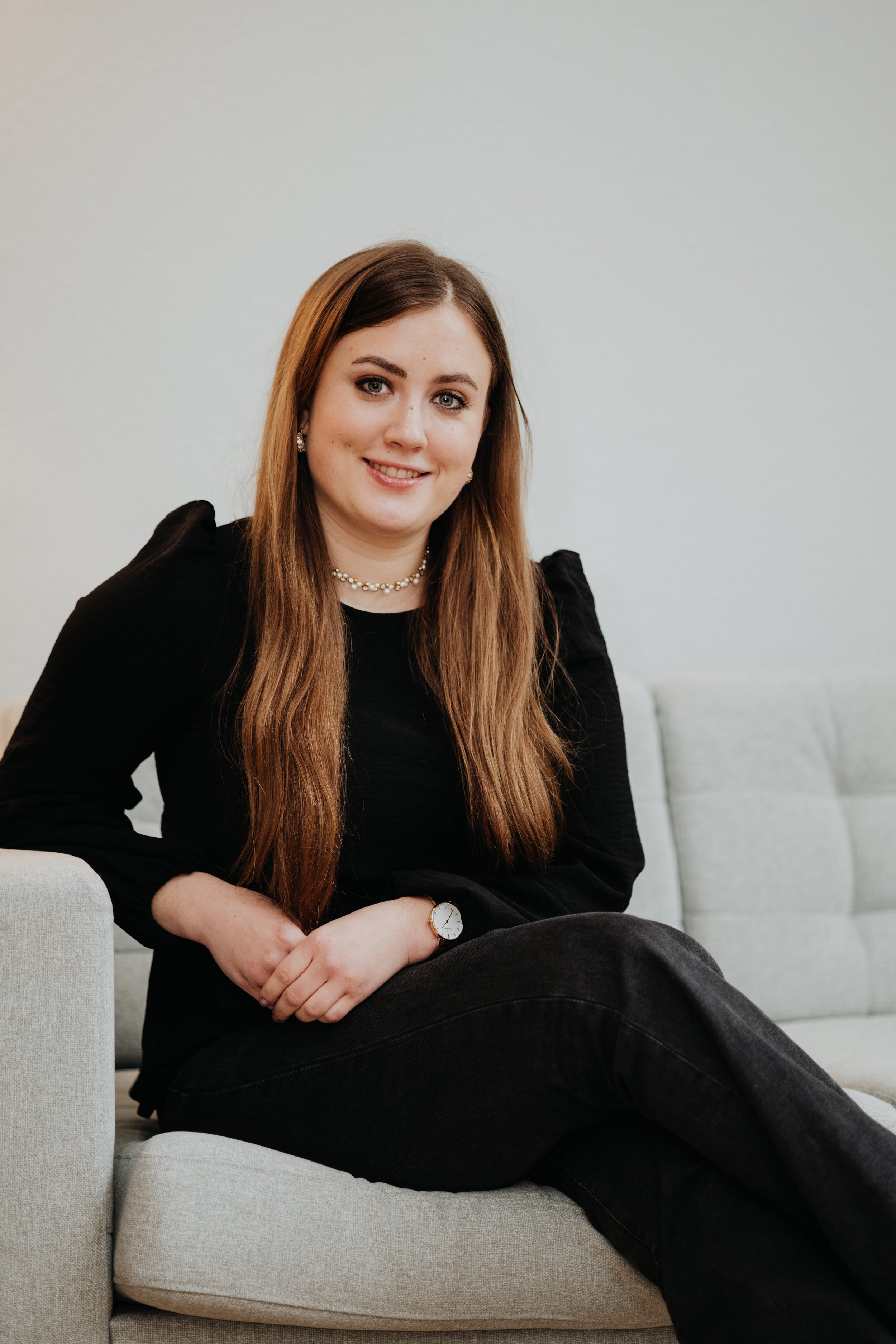 Junge Frau mit langem braunem Haar, schwarzem Oberteil, schwarzen Hose, sitzend auf einem Sofa, lächelnd, mit einer Armbanduhr und Perlenkette, vor hellem Hintergrund.