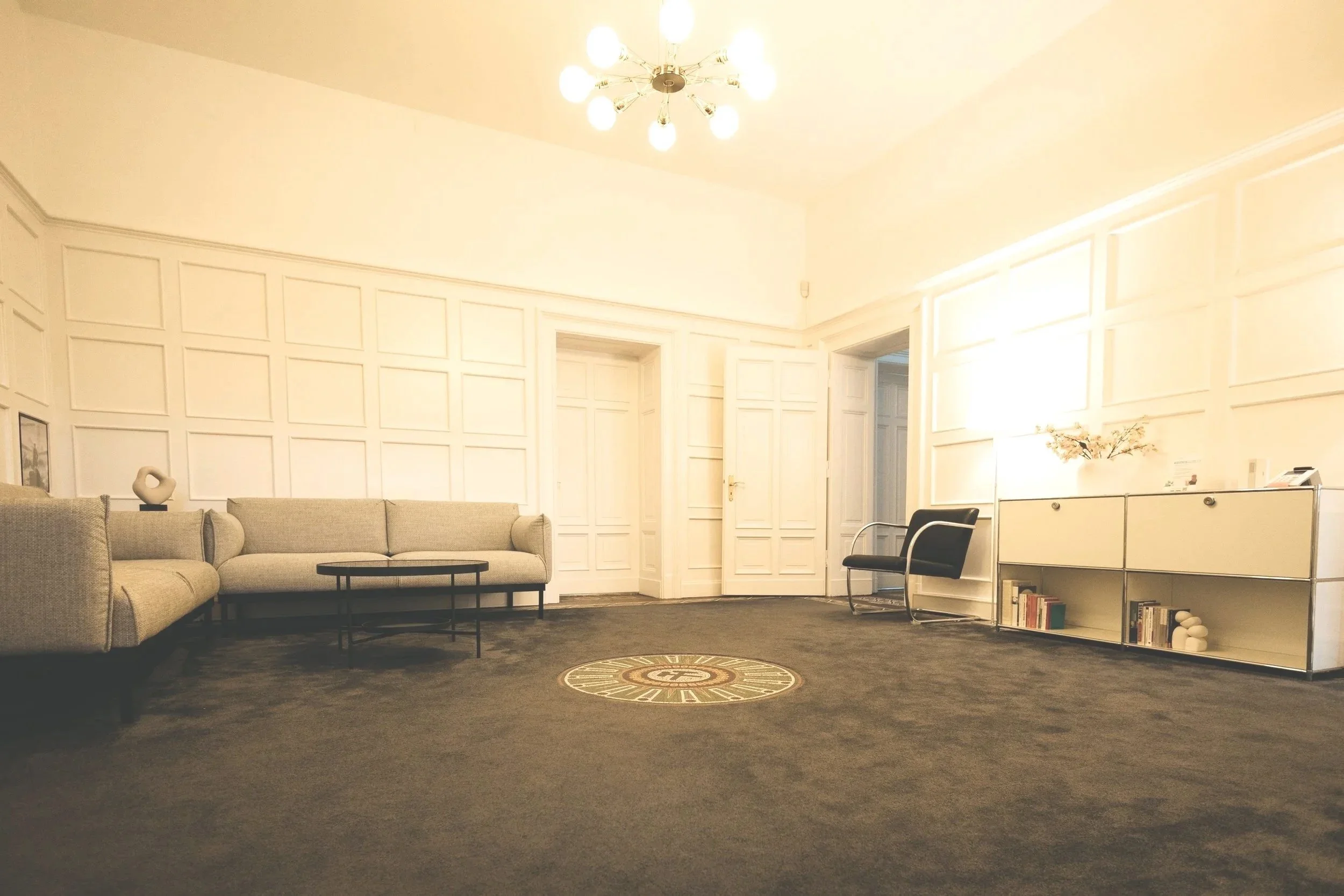 Ein eleganter, heller Sitzraum mit weißen Wänden, beigen Sofas, einem schwarzen Couchtisch, einem schwarzen Sessel und einem weißen Regal mit Büchern. Eine Deckenleuchte mit mehreren Glühbirnen hängt an der Decke.