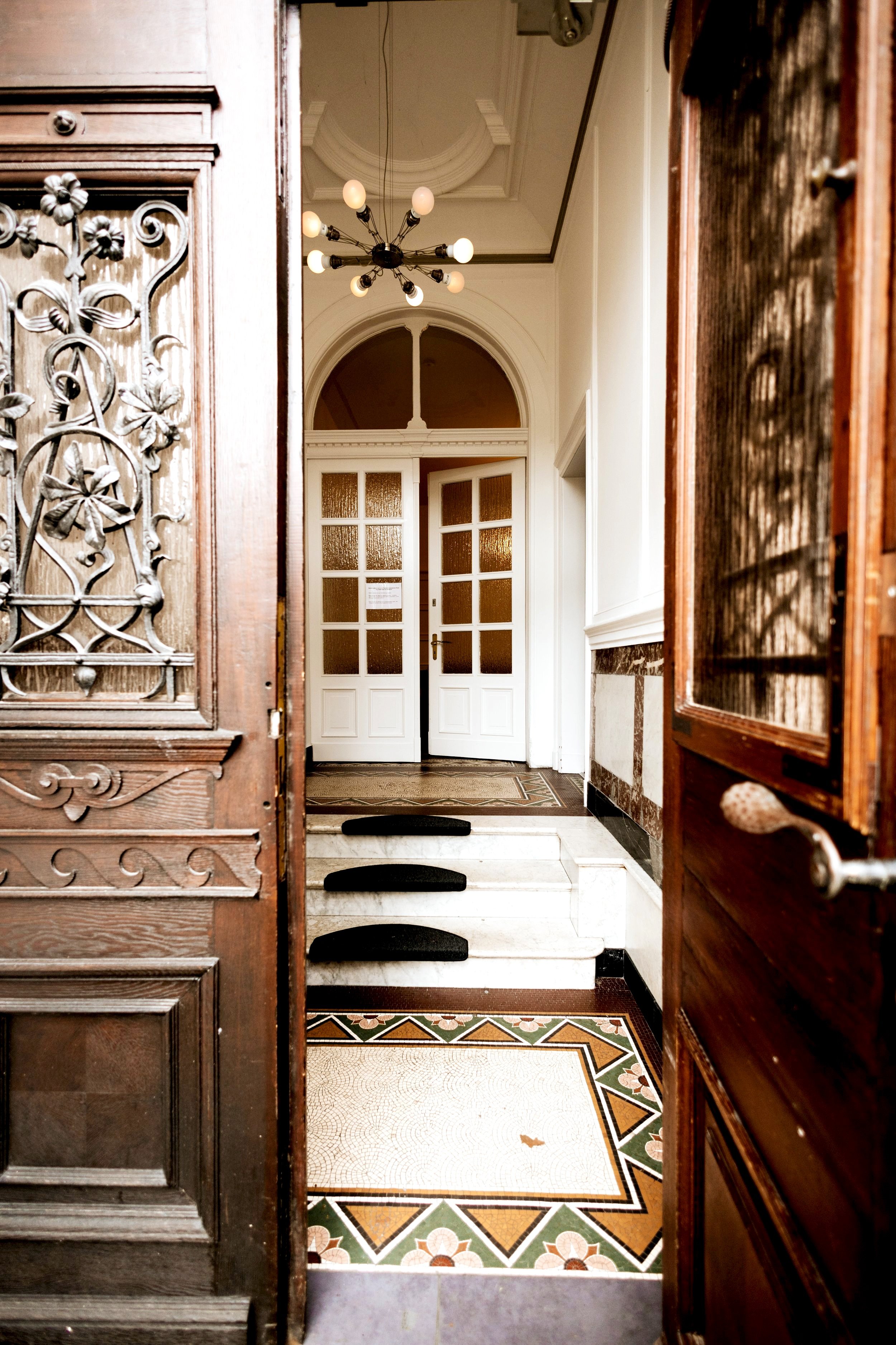 Blick in den Eingangsbereich eines Hauses mit offenen Türen, Flur mit Mosaikfliesen und Treppe, Deckenlampe und große Fenster