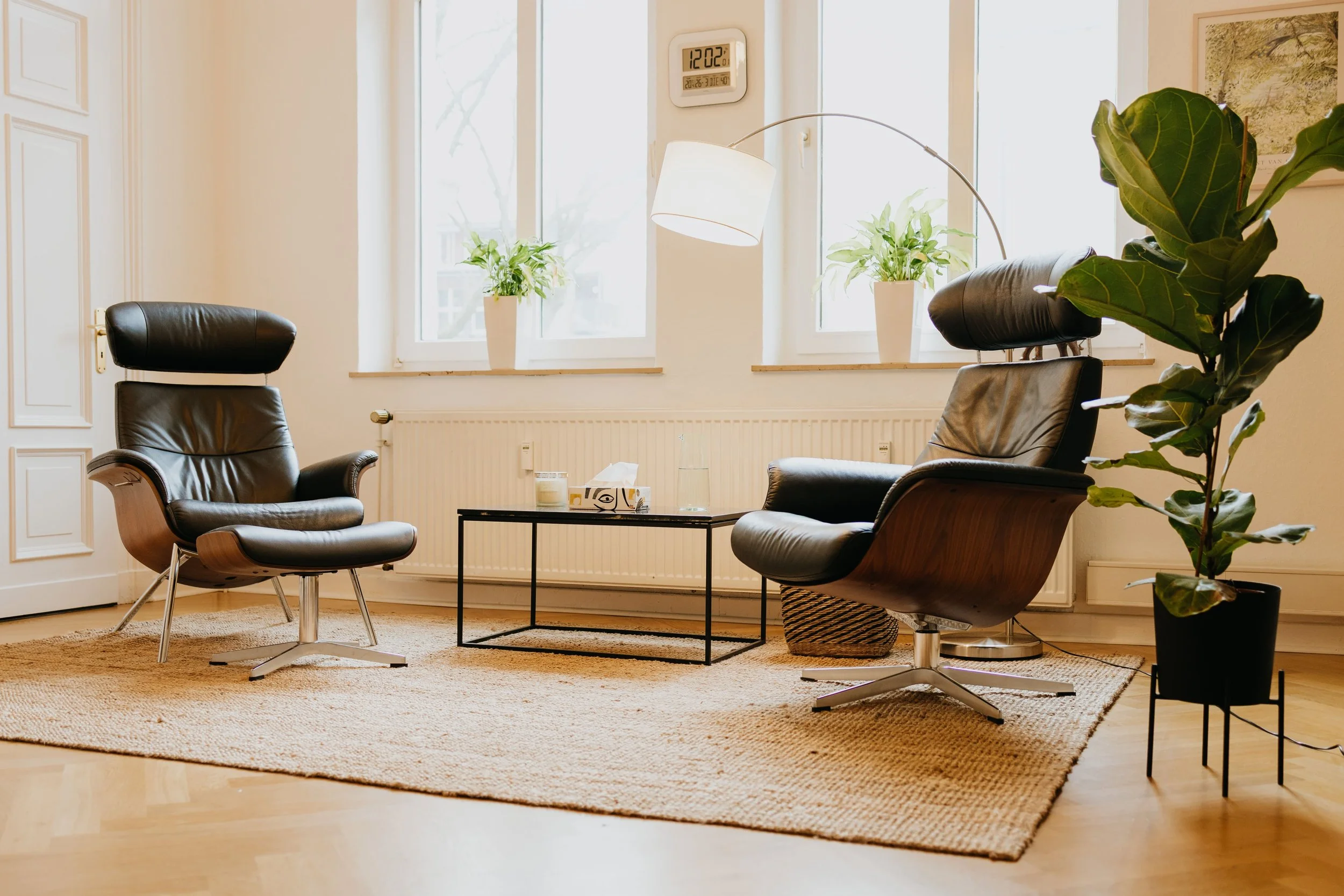 Zwei schwarze Ledersessel in einem Wohnzimmer mit Fenster und Pflanzen, Couchtisch mit Kerze und Wasserflasche, Teelicht, Teppich auf Holzboden.