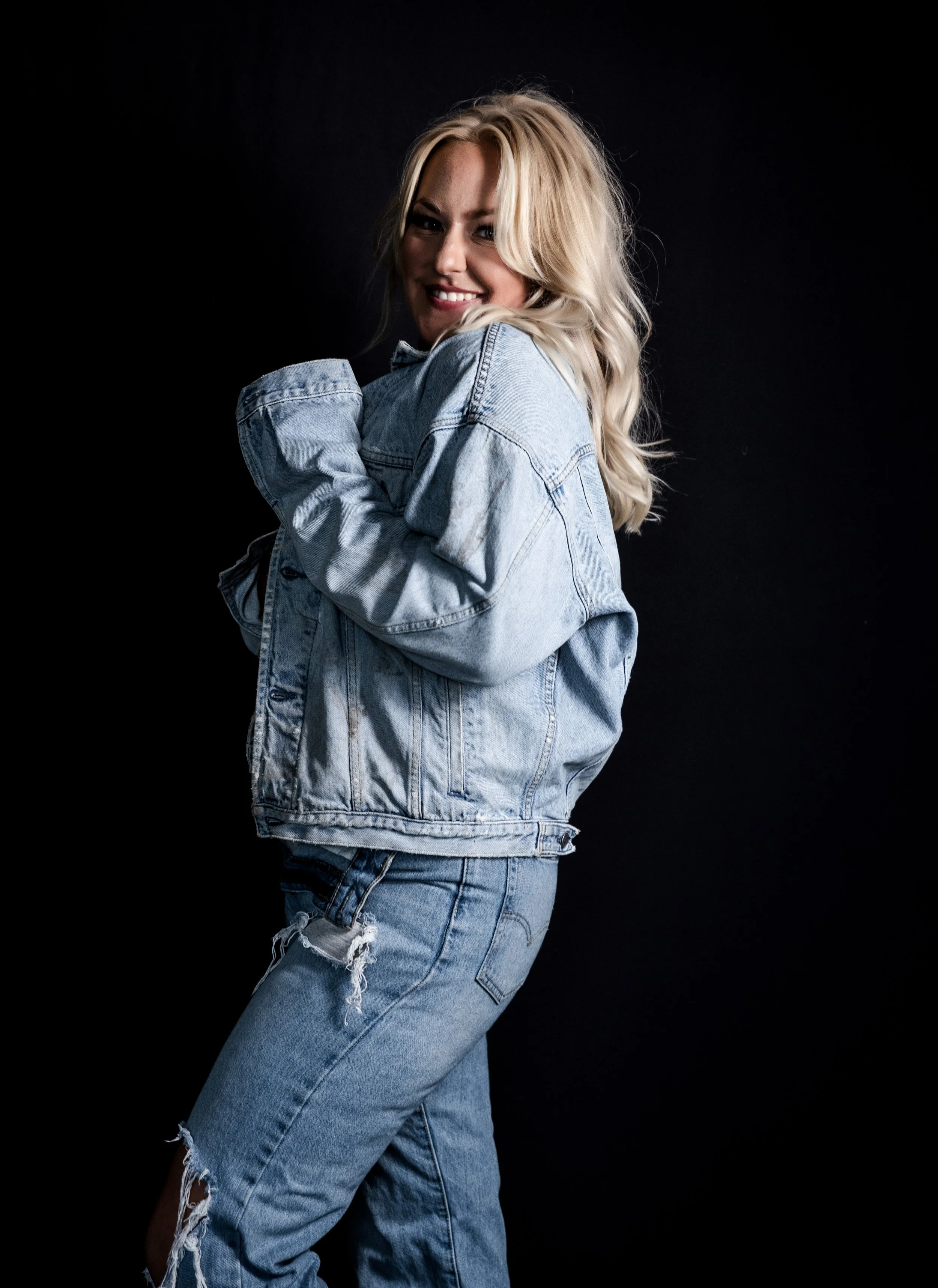 Woman in denim jacket and ripped jeans against dark background.