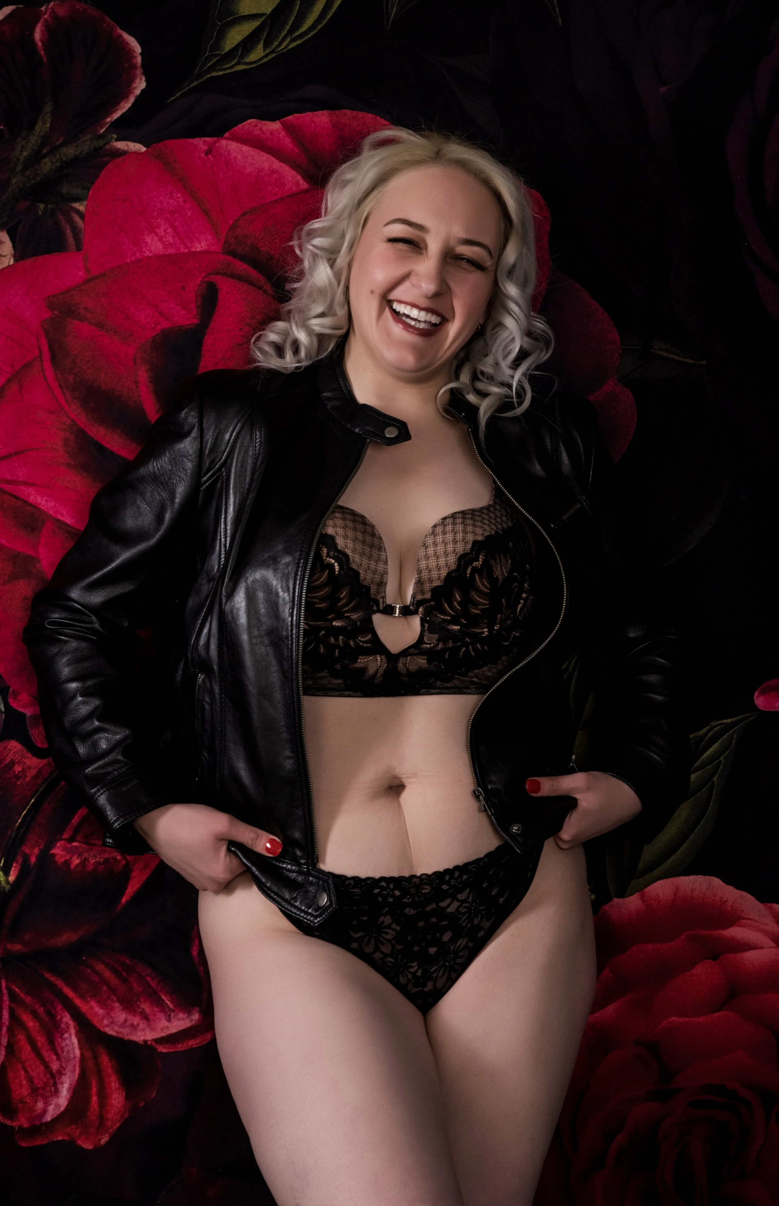 Woman in black lingerie and leather jacket smiling in front of floral background.