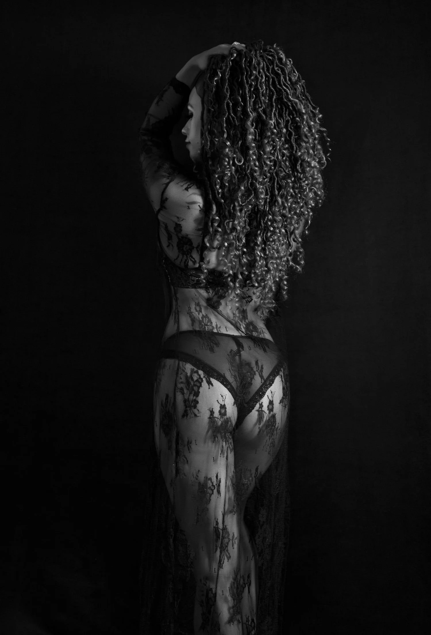 Black and white photo of a woman in lace lingerie, with long curly hair, standing in profile against a dark background.