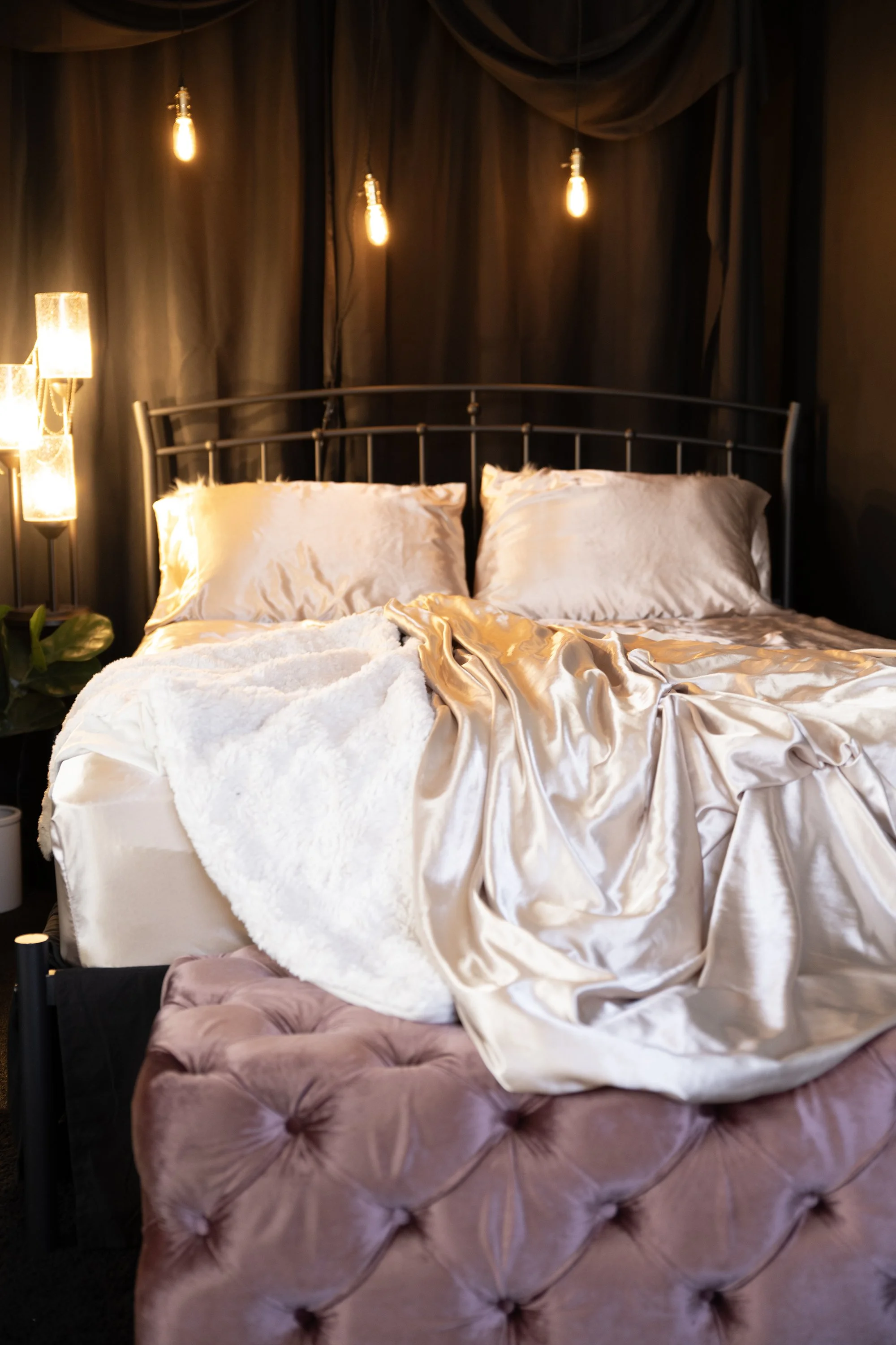 A bedroom with a bed, satin sheets, a tufted pink ottoman at the foot of the bed, dark curtains, and hanging Edison-style bulbs warm lighting.