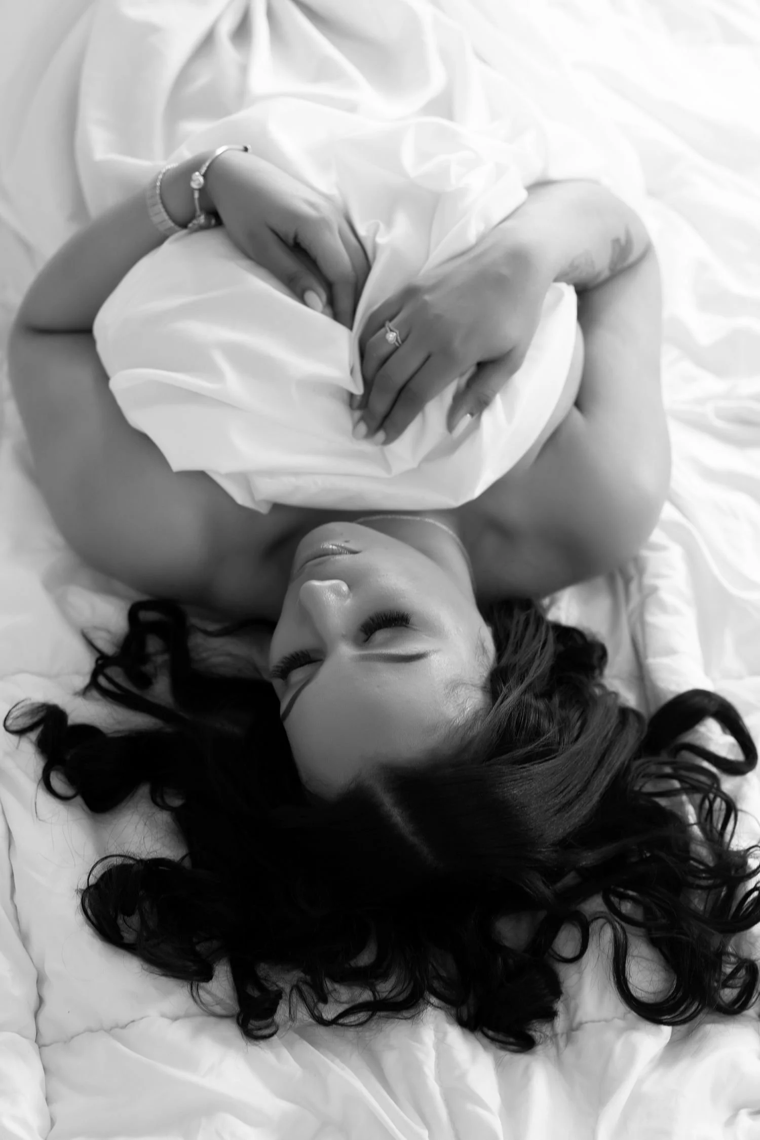 Black and white photo of a woman lying on a bed with tousled hair, draped in a white sheet, holding it with her hands.