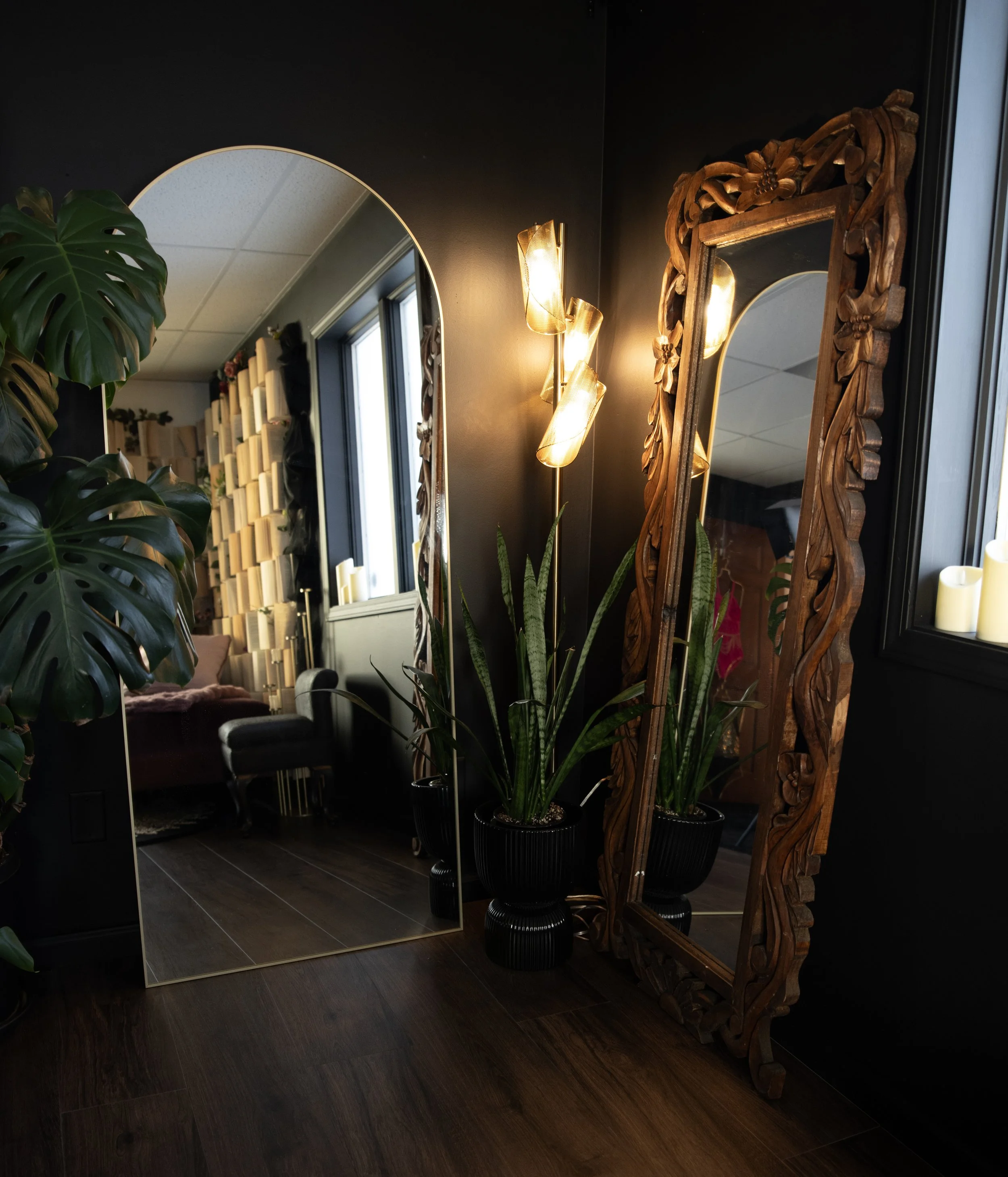 A room with dark walls and wooden flooring, featuring two tall mirrors with one being adorned with intricate wooden carvings, a large green plant, a standing lamp with three glass shades, and a window letting in natural light.