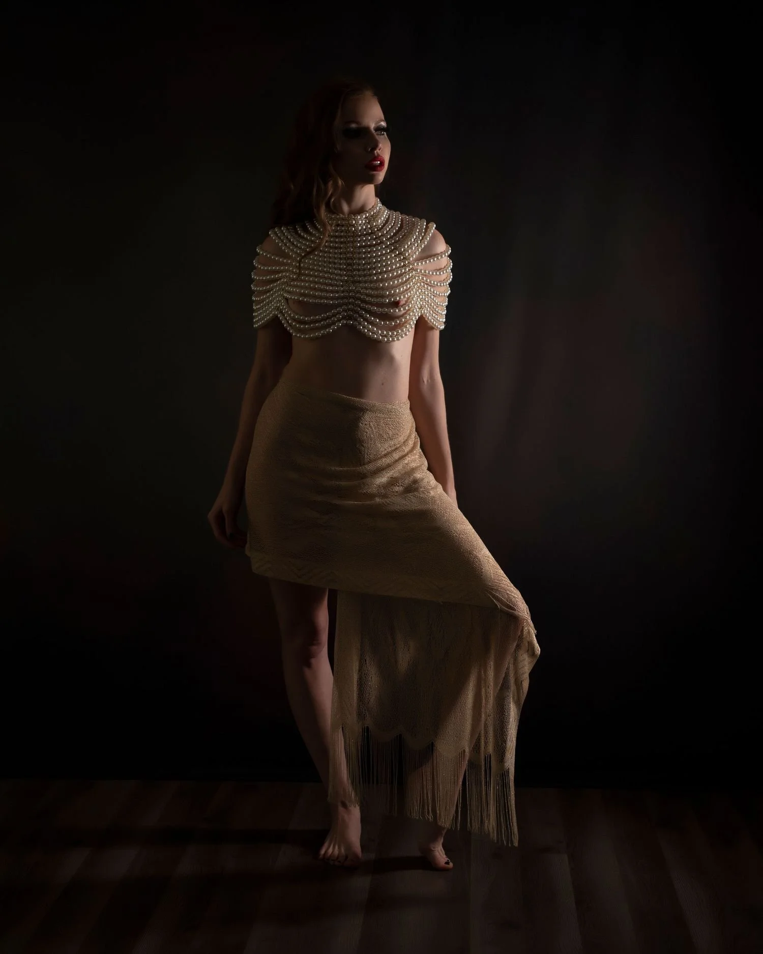 Woman wearing a layered pearl top and beige skirt, posing in a dark setting.