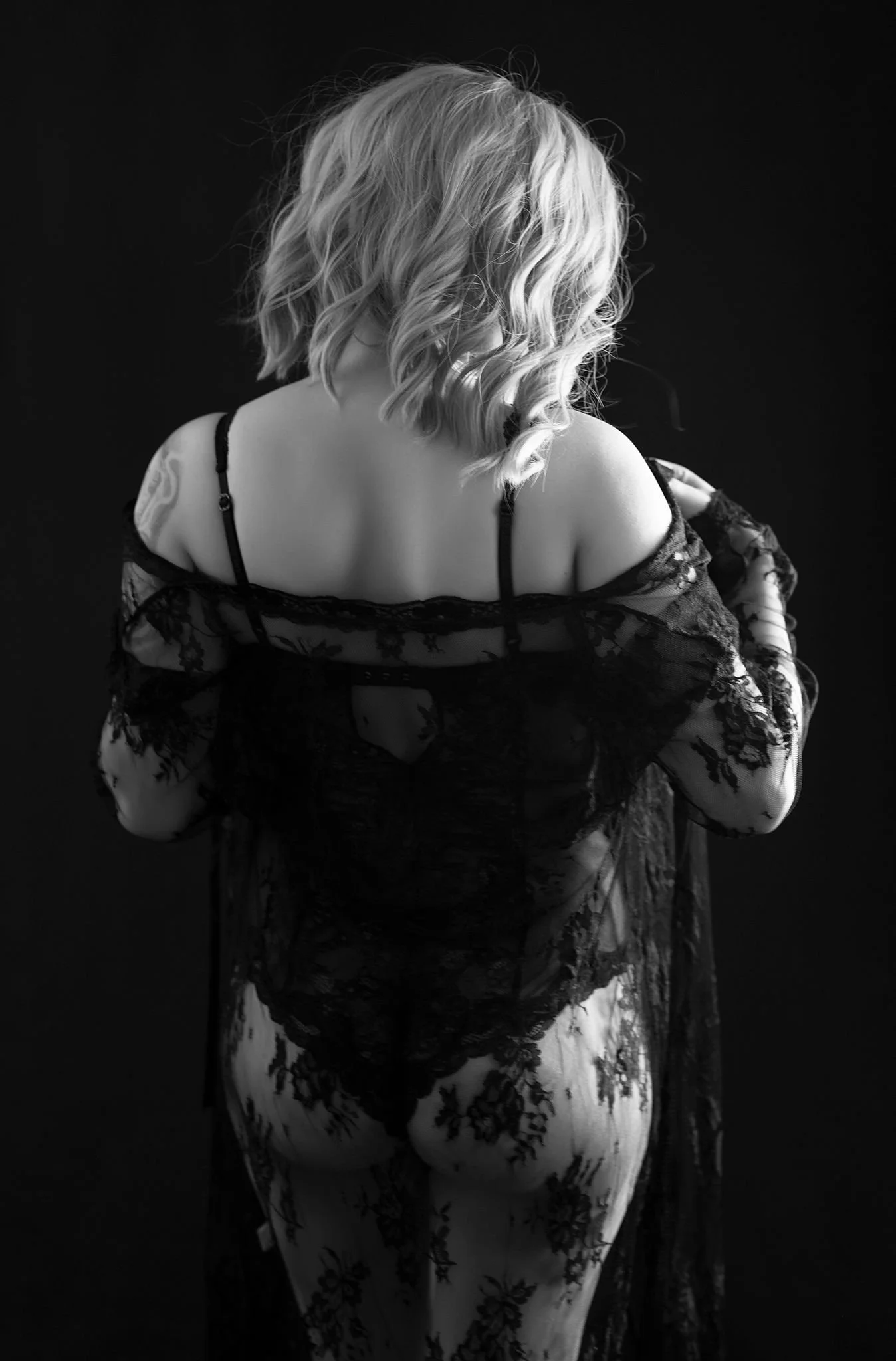 Rear view of a woman with shoulder-length hair wearing black lace lingerie against a dark background.