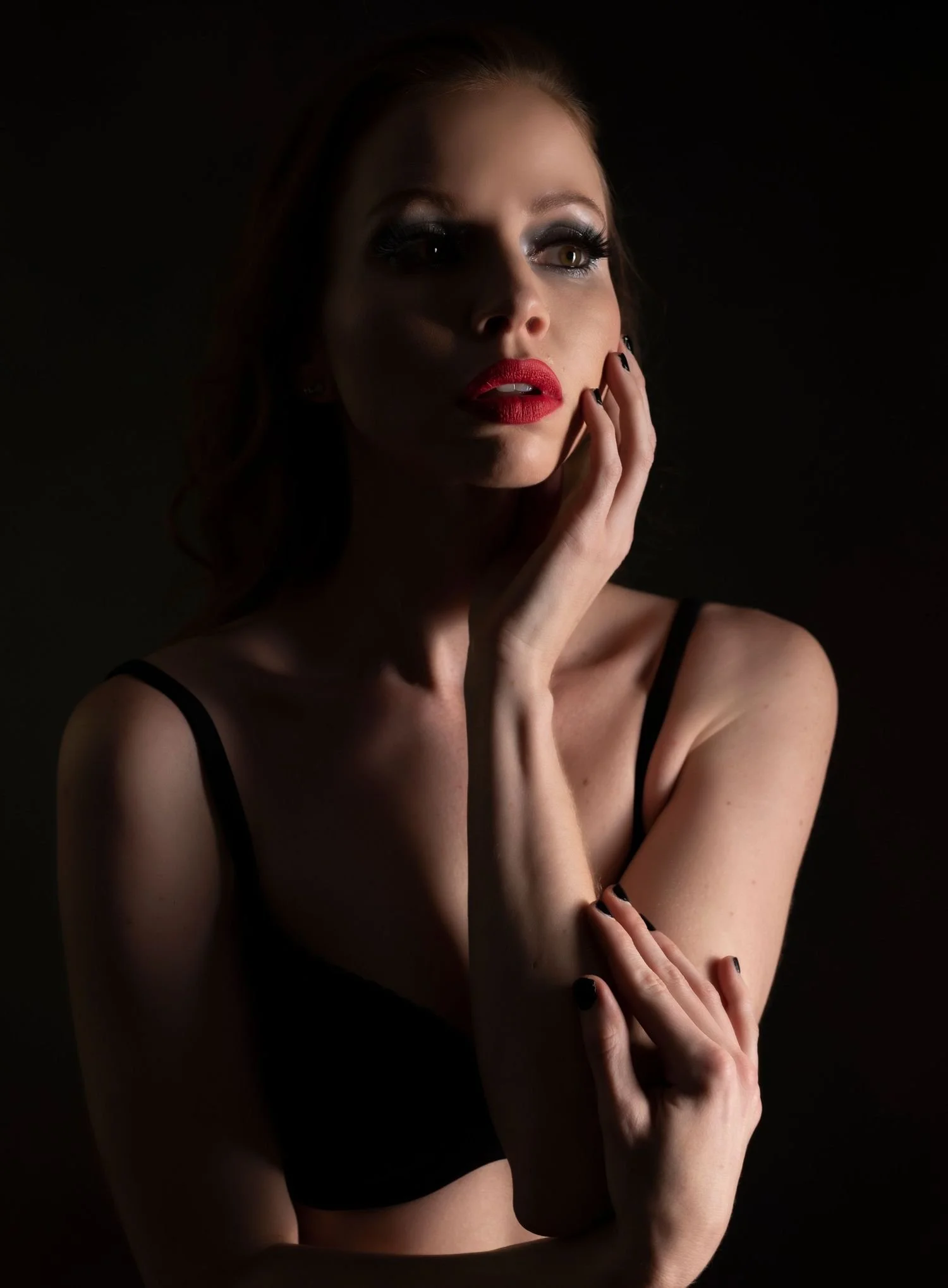 A woman in dramatic low-key lighting, wearing red lipstick and a black top, with a hand on her face.