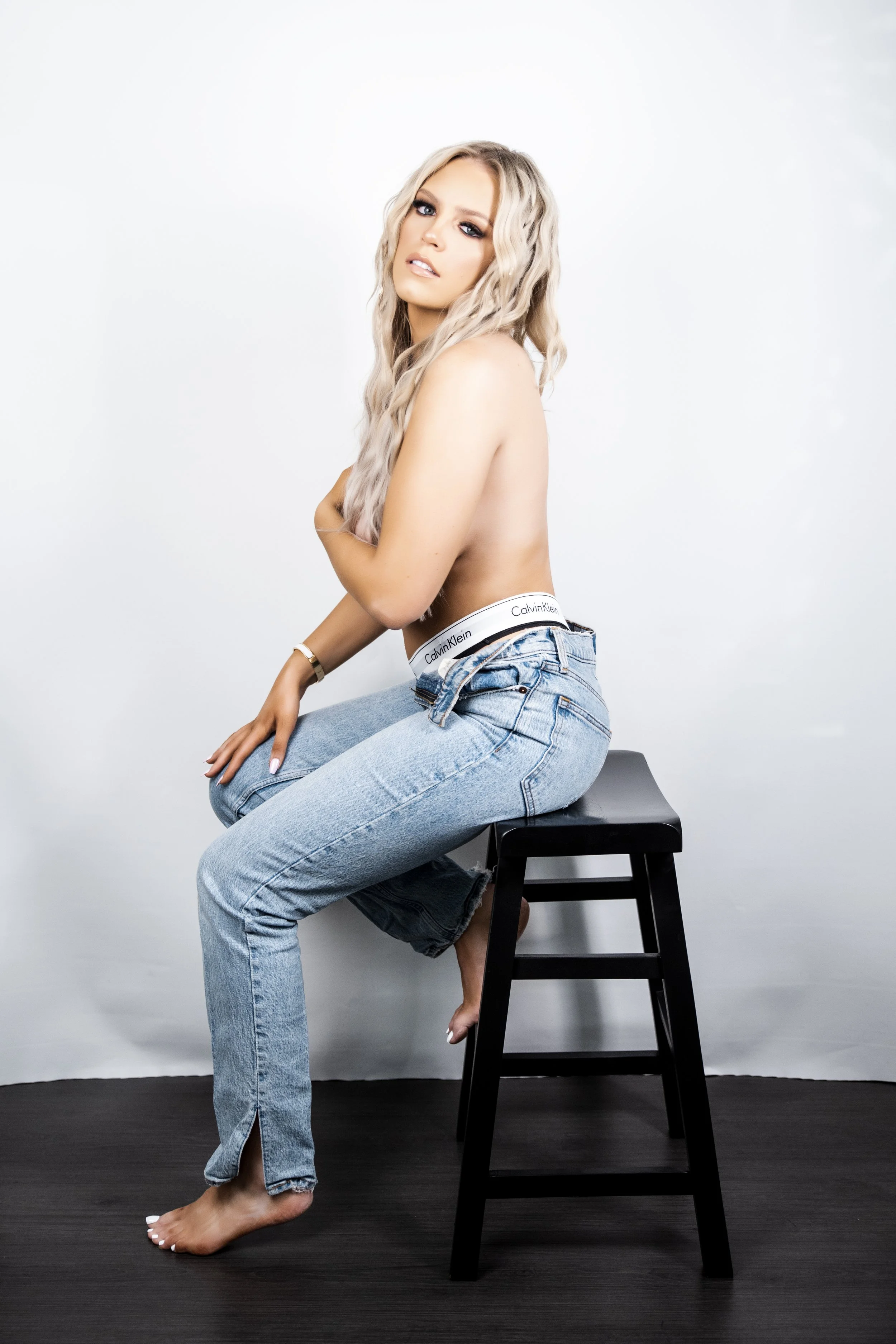 Person with long blonde hair wearing jeans sitting on a stool against a white background.