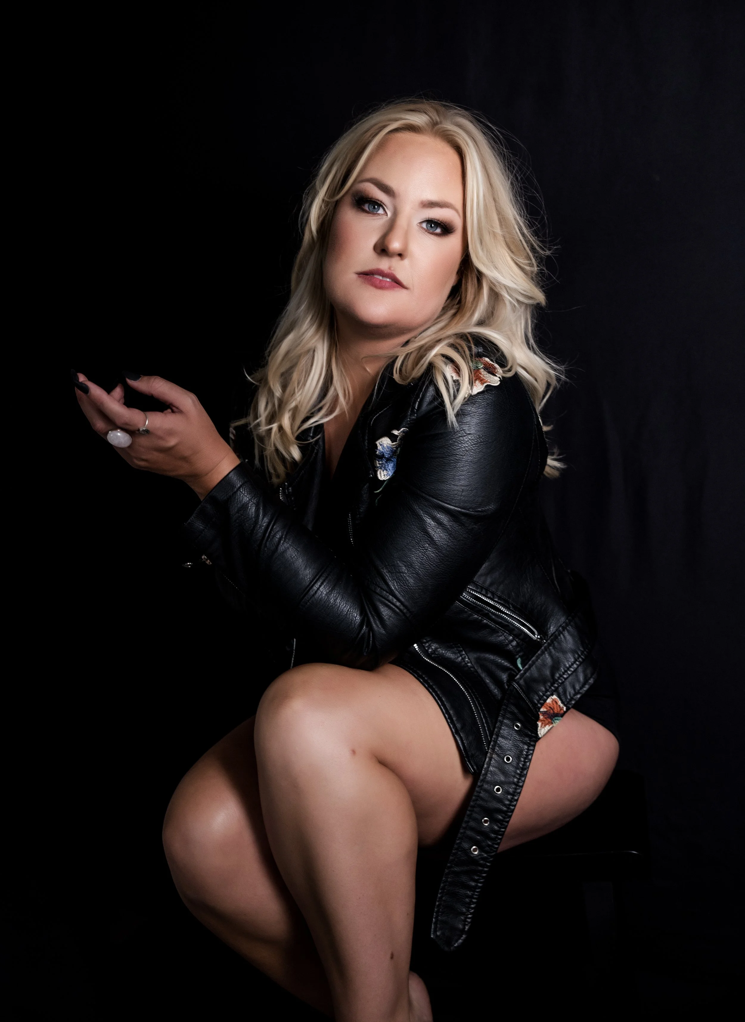 Woman with long blonde hair, wearing a black leather jacket, sitting on a stool against a dark background.