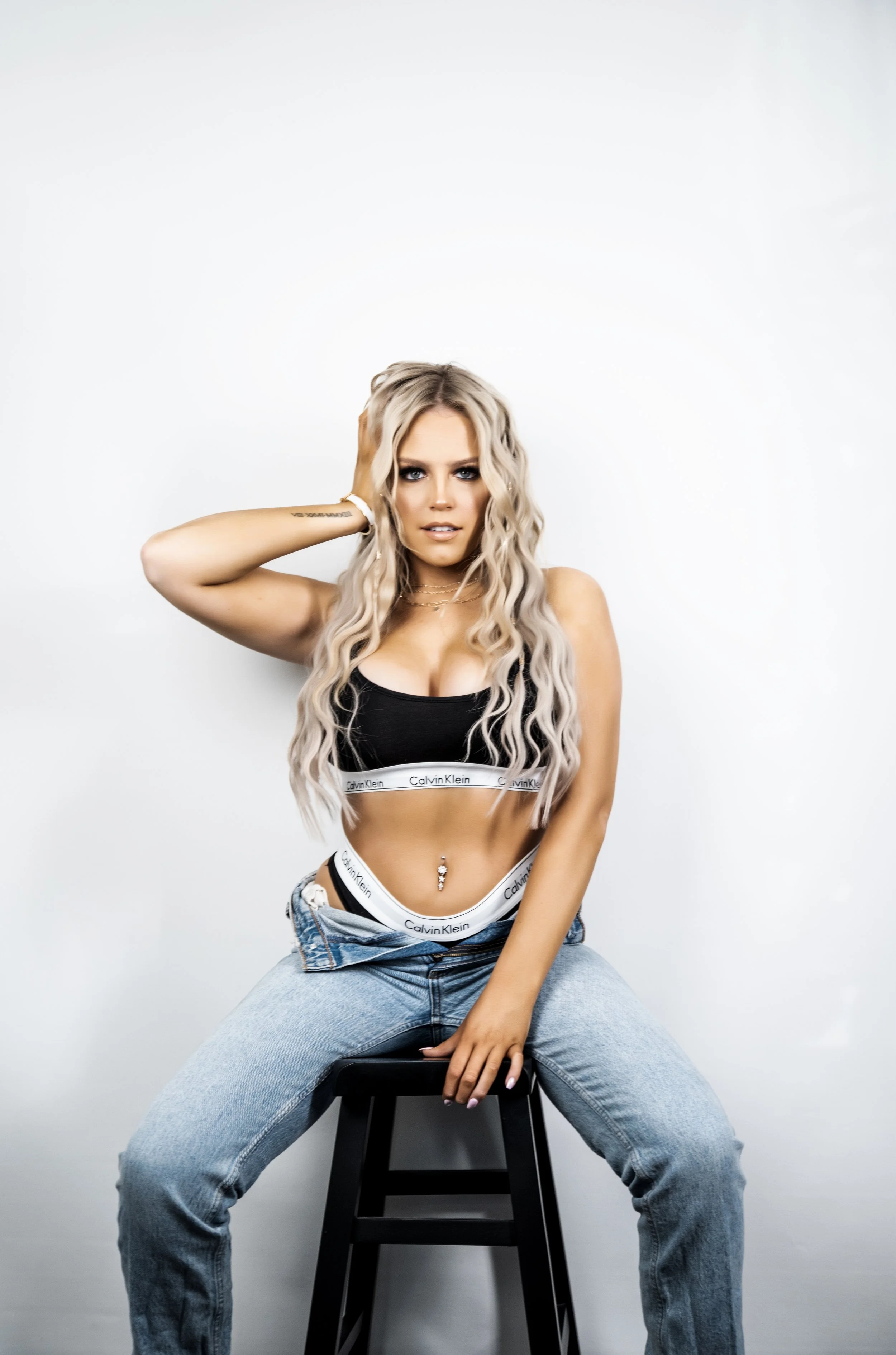 A person with long blonde hair, wearing a Calvin Klein crop top and jeans, sits on a stool in front of a white background.