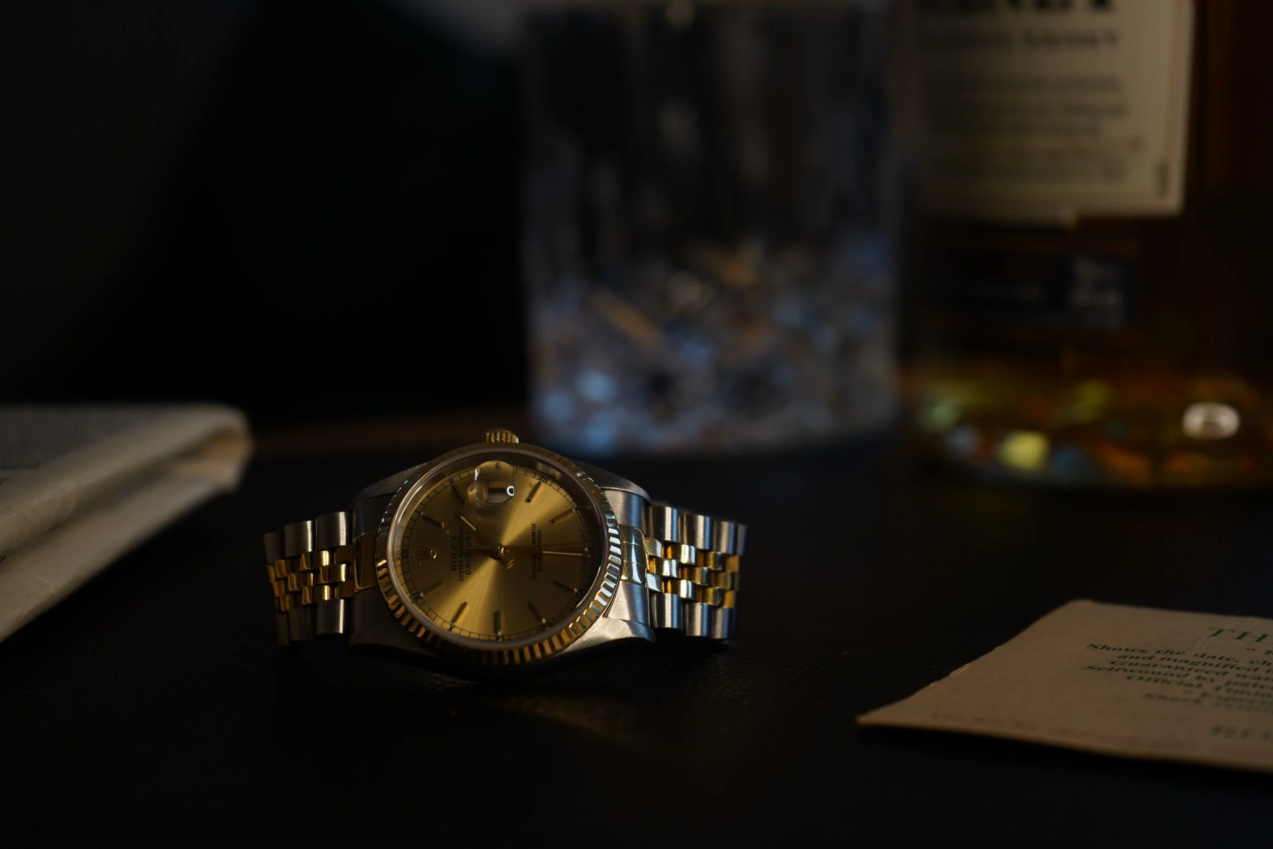 Et Rolex ur med guld- og sølvrende, placeret på en sort overflade, omgivet af papirer og en glasbeholder i baggrunden.