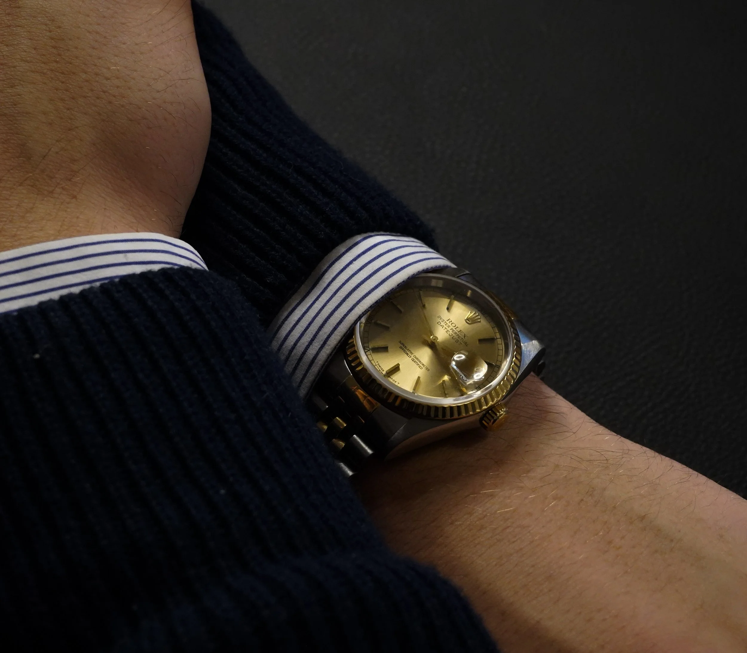 Person iført en gulddialet Rolex-ure med stålrem, iført en stribet skjorte og en mørk trøje.
