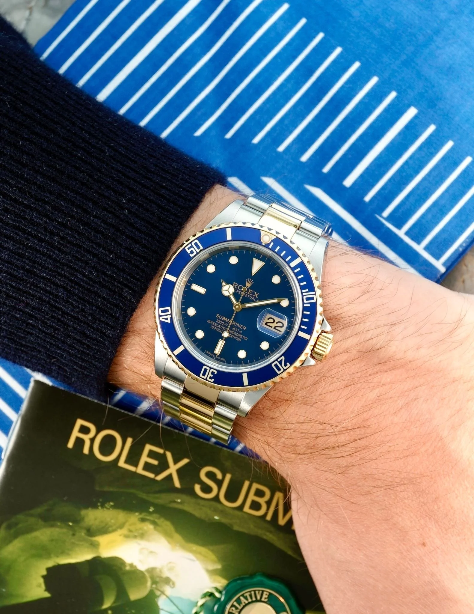Et håndled med en Rolex Submariner ur i blå og guld, et sted hvor en person holder en brochure med teksten «ROLEX SUBMARINER».