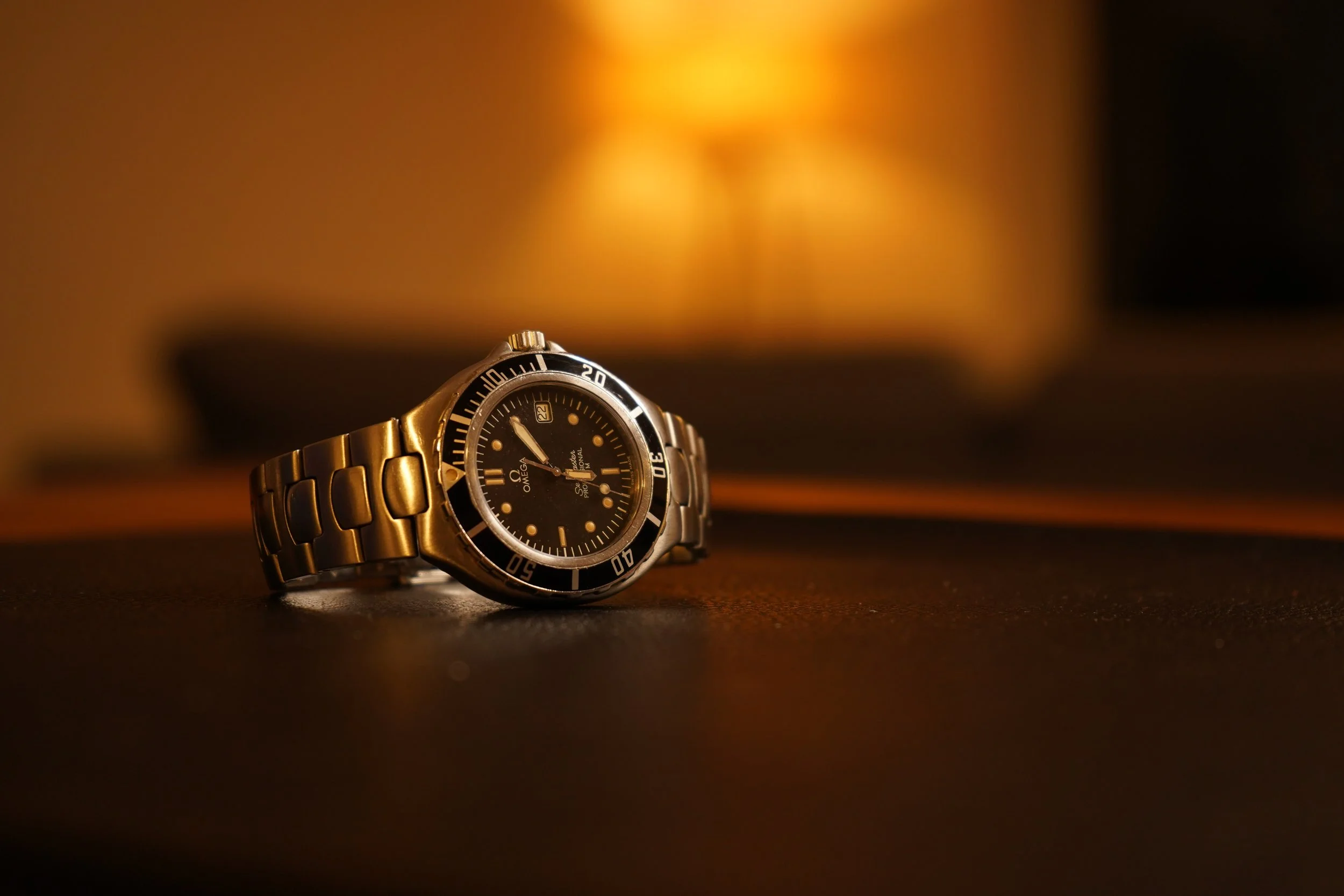Et Omega Seamaster 200M med sort ansigt og metal rem lagt på en mørk overflade, med en varm, uklar lampe i baggrunden.