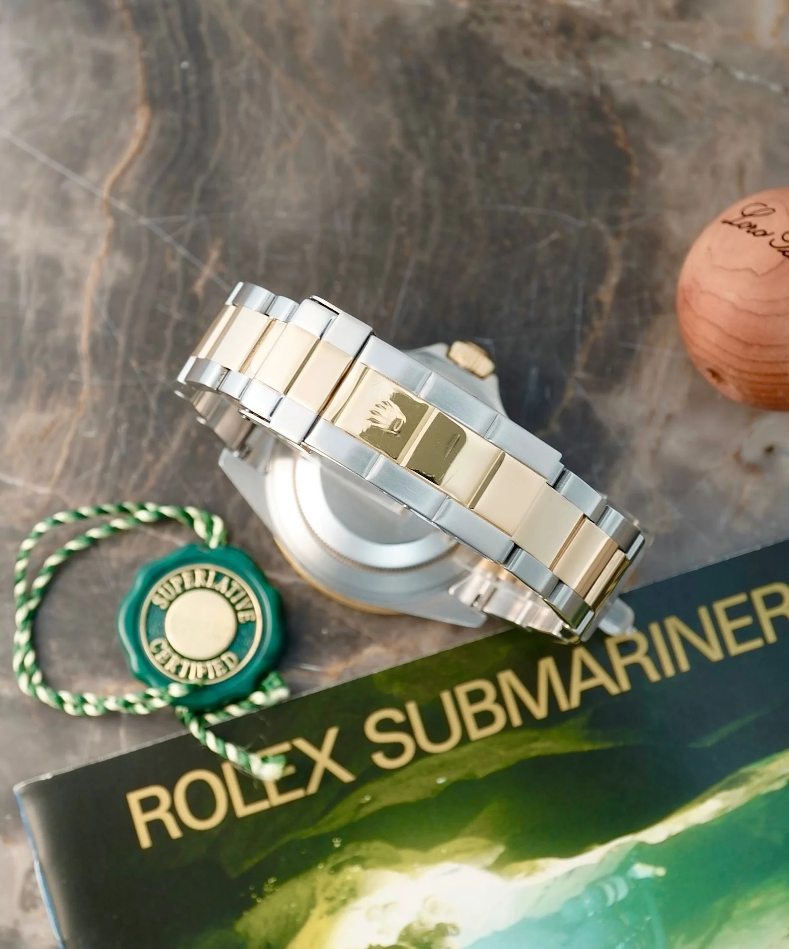 En Rolex Submariner ur med guldbånd og en grøn og guld tag, placeret på en bordflade sammen med en bog om Rolex Submariner og en trææske.