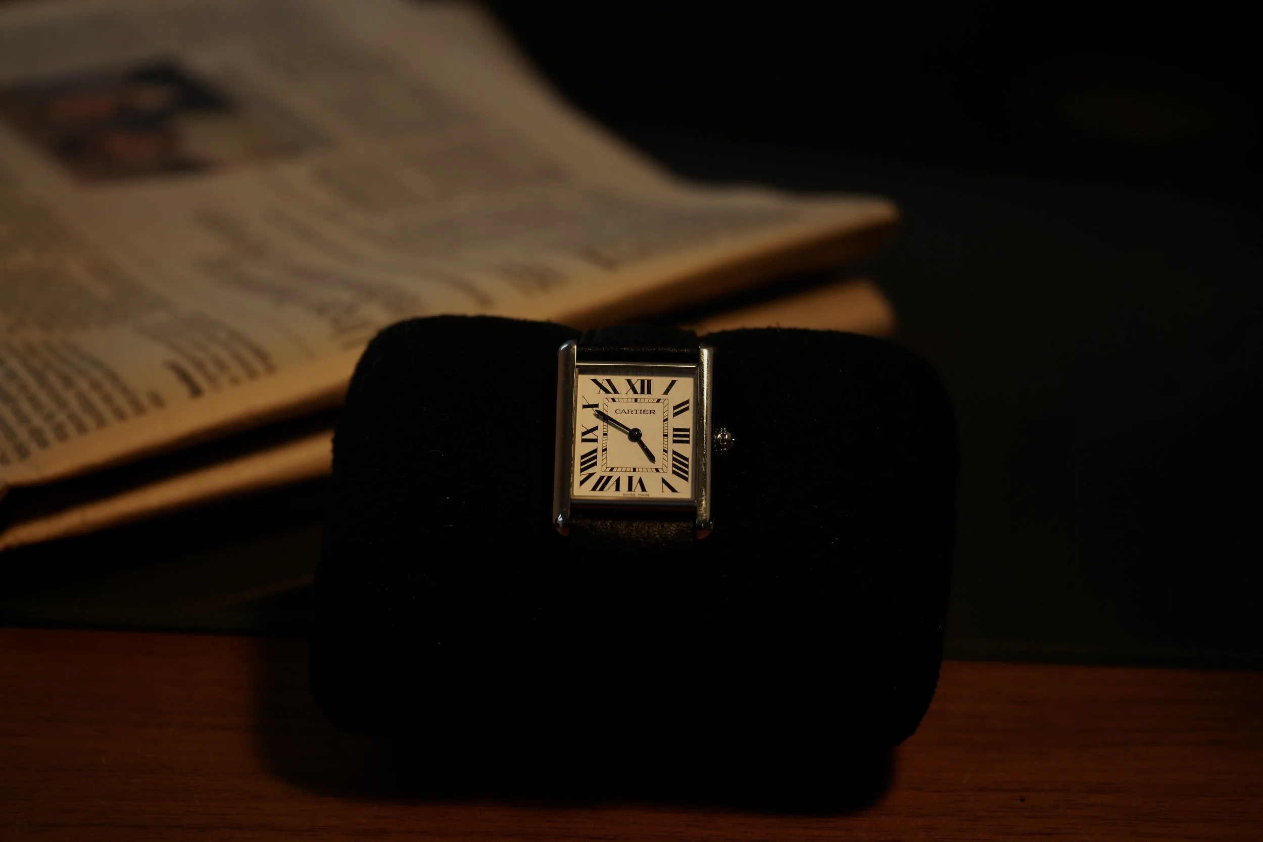 Et Cartier tank med hvid urskive, sort romertal og sort visere, placeret på en sort pude foran en åben avis, på en træoverflade.