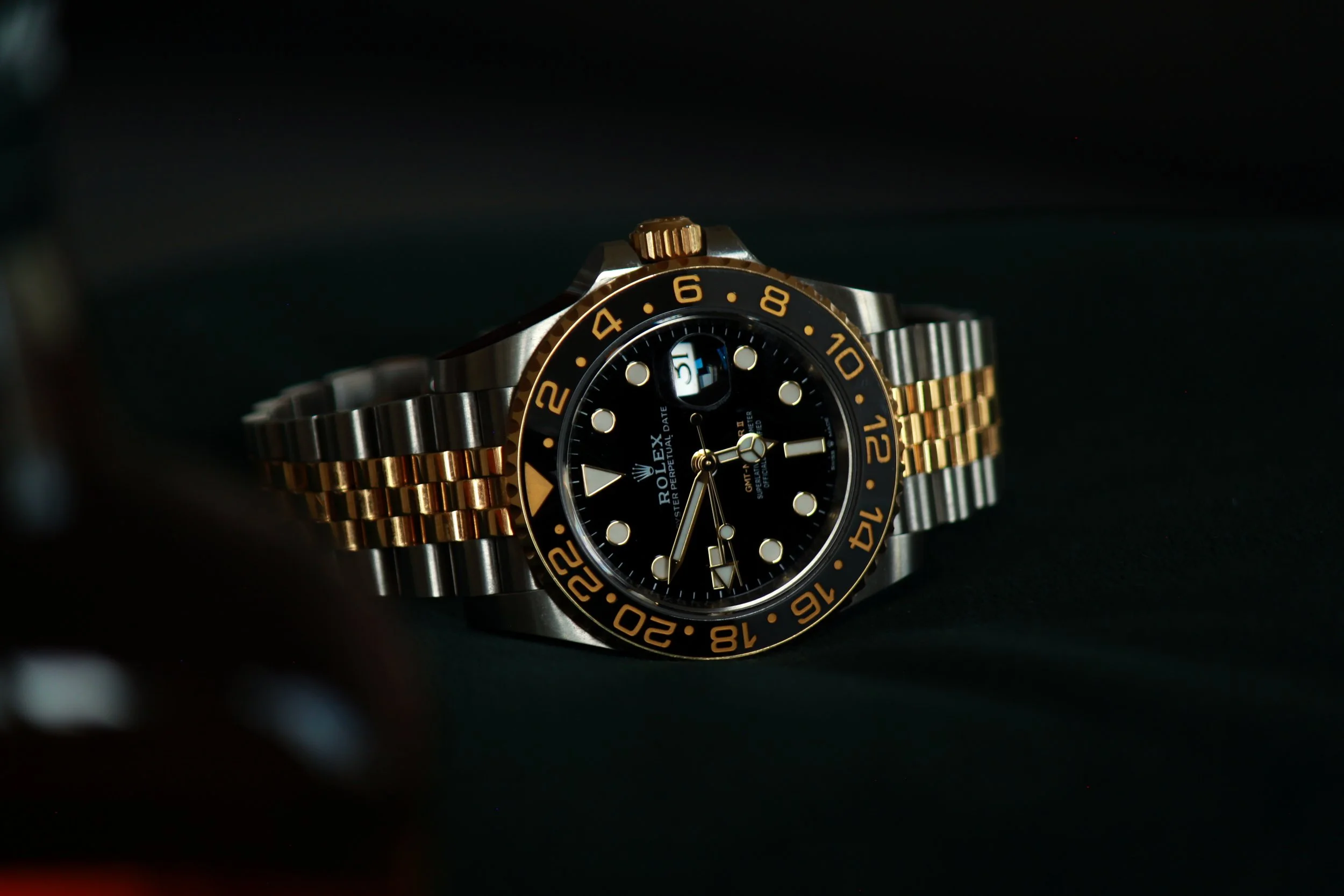 Et Rolex ur med sort urskive, guldbelagt krans, og to-tone metalrem i stål og guld, liggende på en mørk overflade.