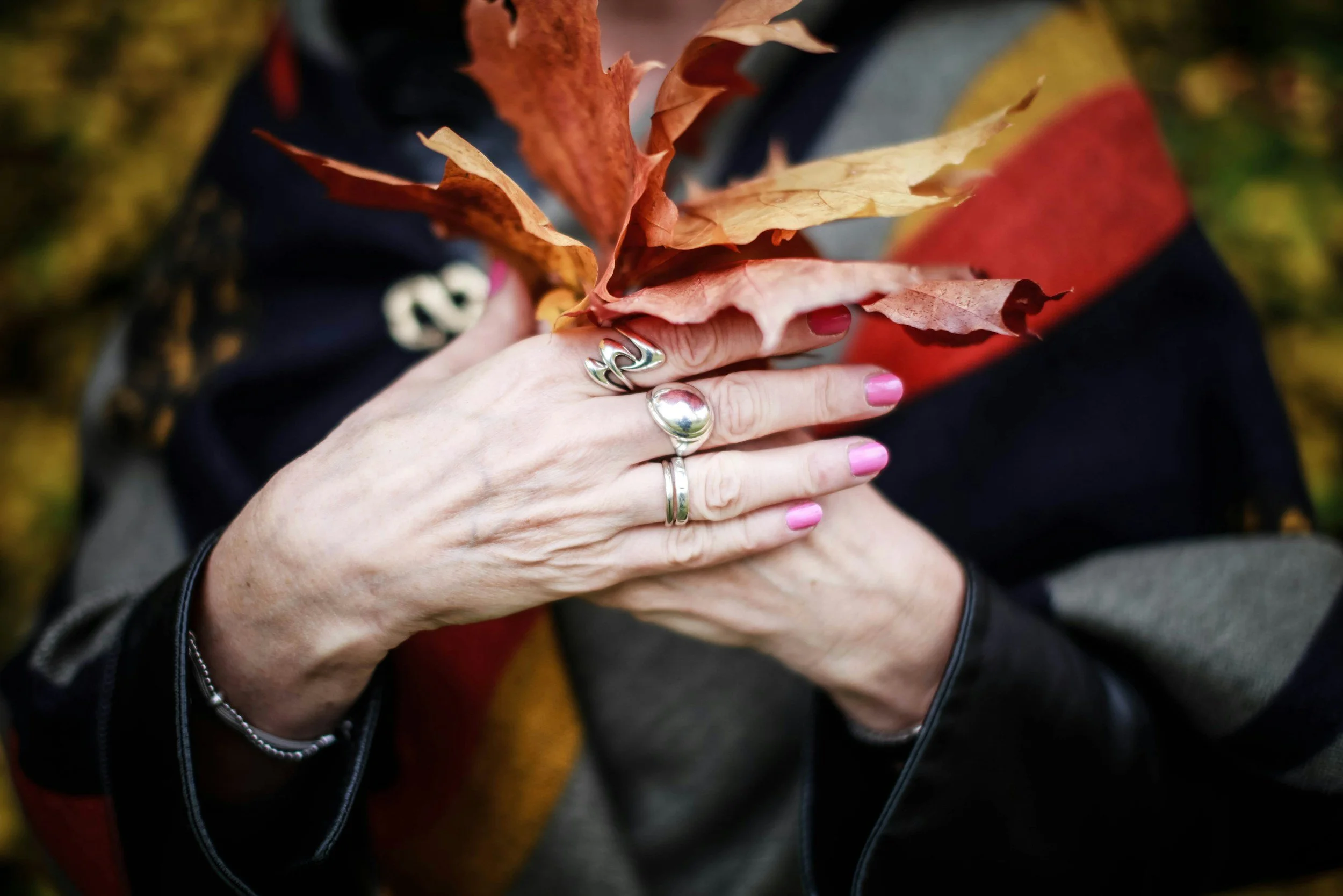 Person holding colorful autumn leaves with rings on fingers, wearing a black jacket and pink nail polish.